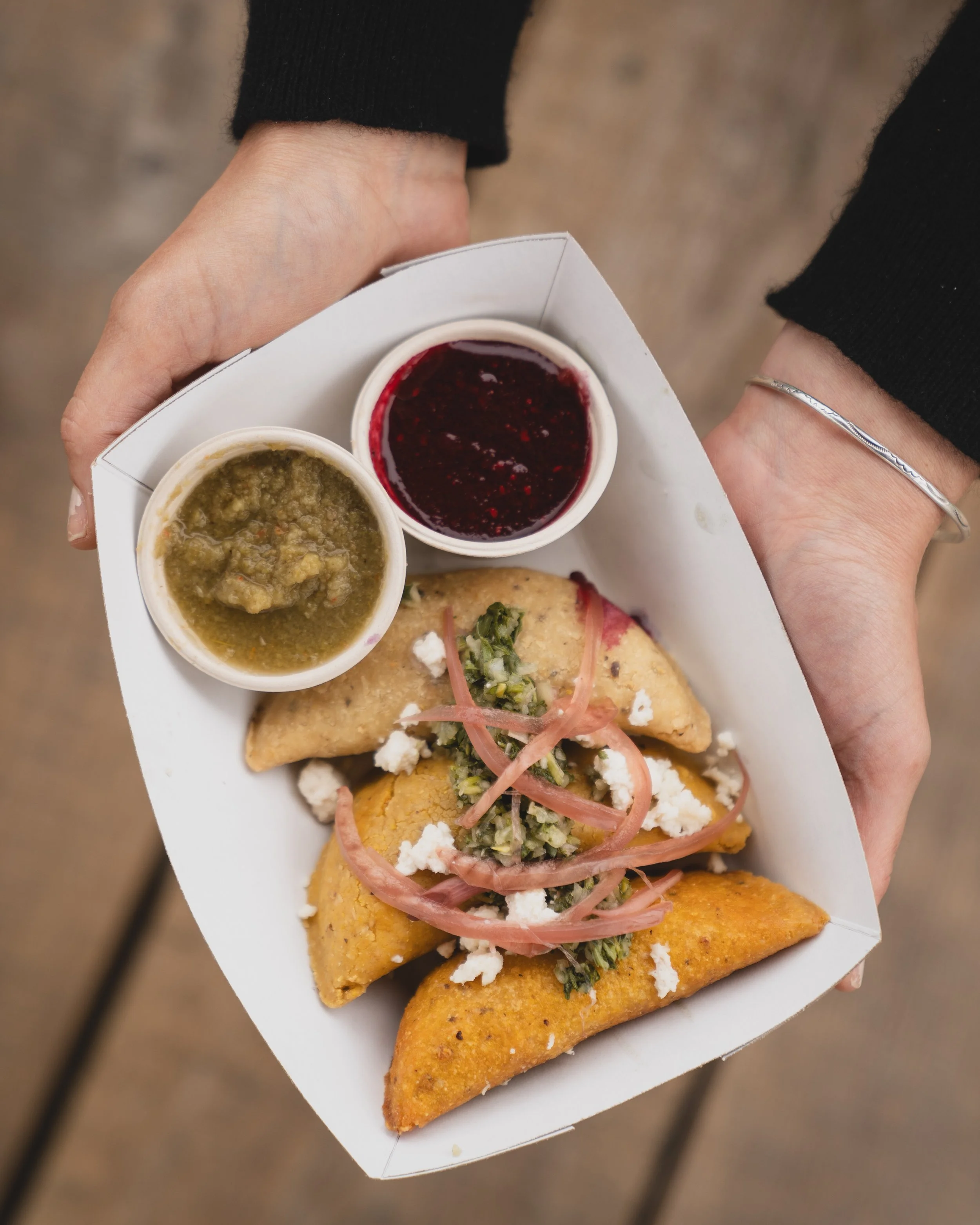 Person holding a white tray with two cups of salsa, chicken and cheese filled empanadas topped with chopped vegetables and pickled onions.