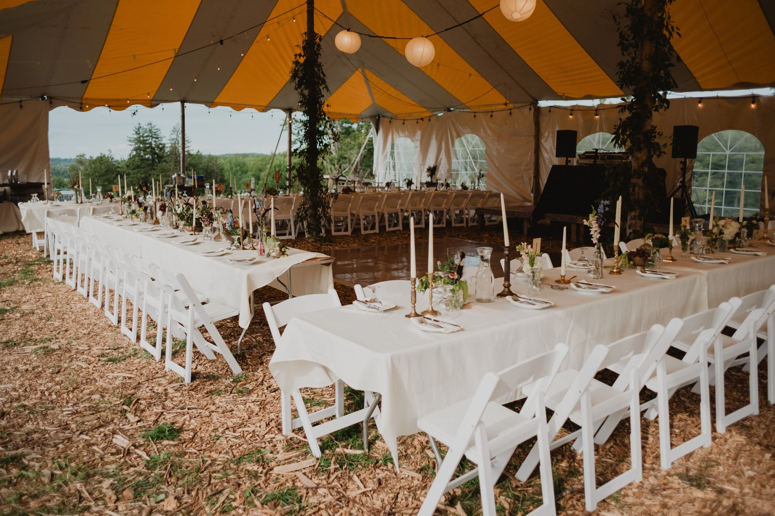 Decorated yellow and white outdoor event tent with long tables covered in white tablecloths, set with plates, glasses, and silverware, surrounded by white folding chairs, with floral centerpieces and candlesticks, overlooking a green landscape.