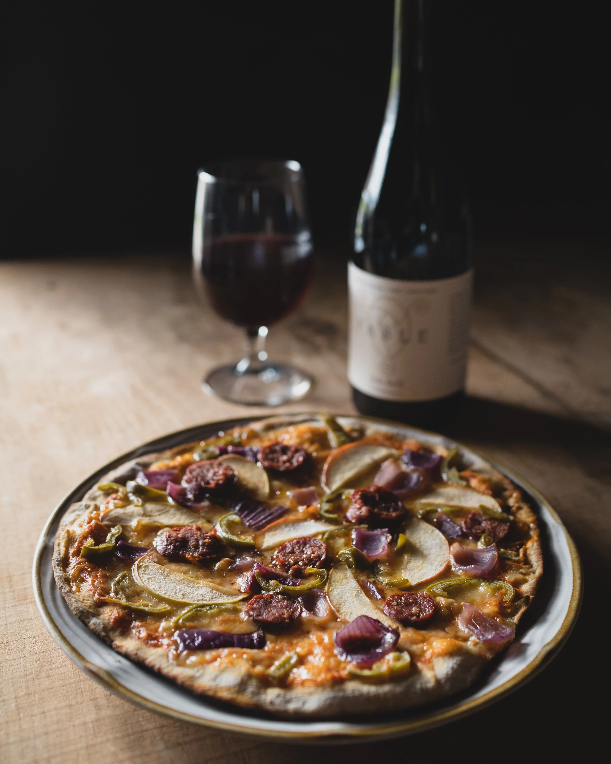 a bottle of wine and a glass of wine paired with a flatbread pizza made with meat and veggies from our farms