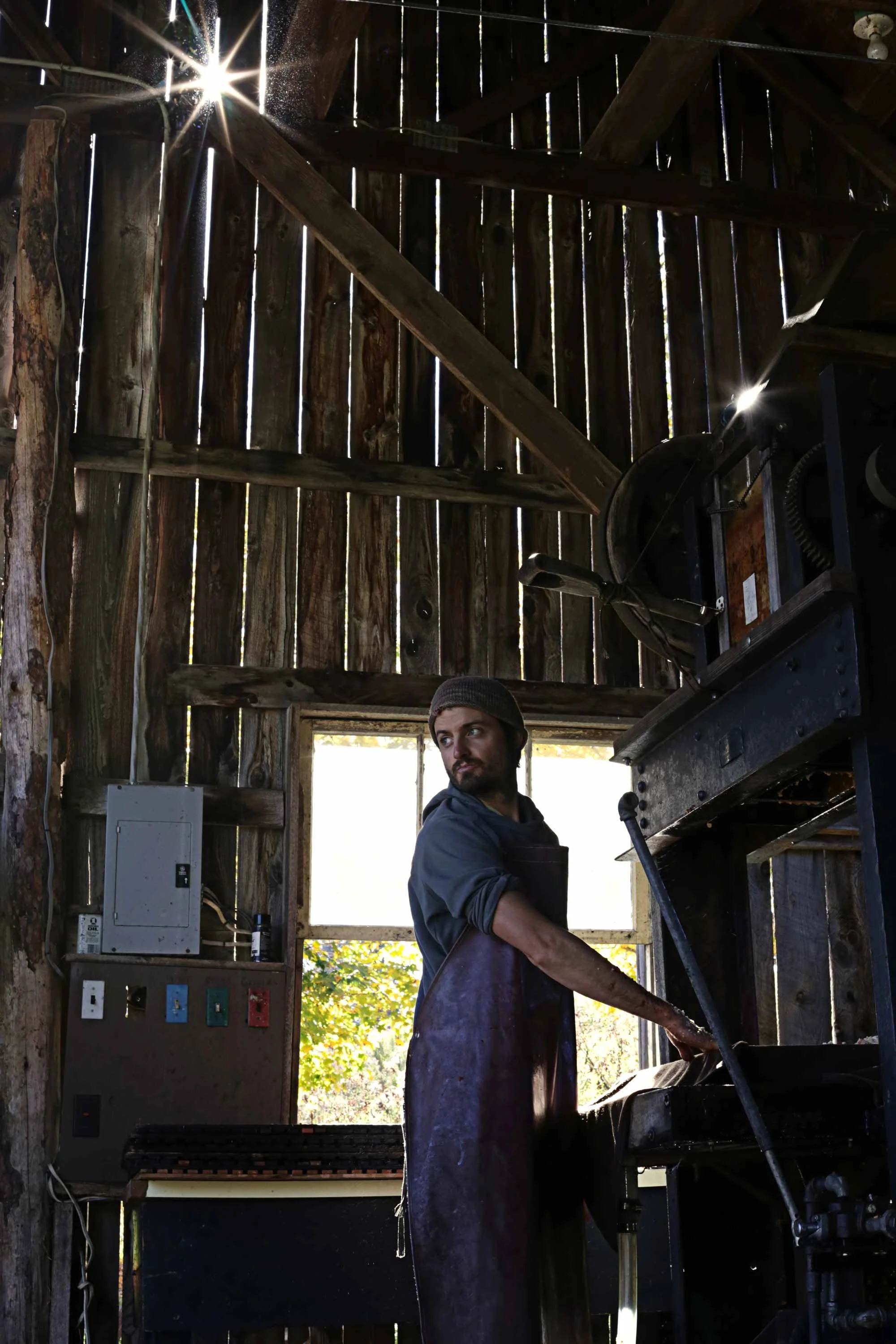 A man working with a machine inside a rustic wooden shed, sunlight streaming through gaps in the wooden walls.
