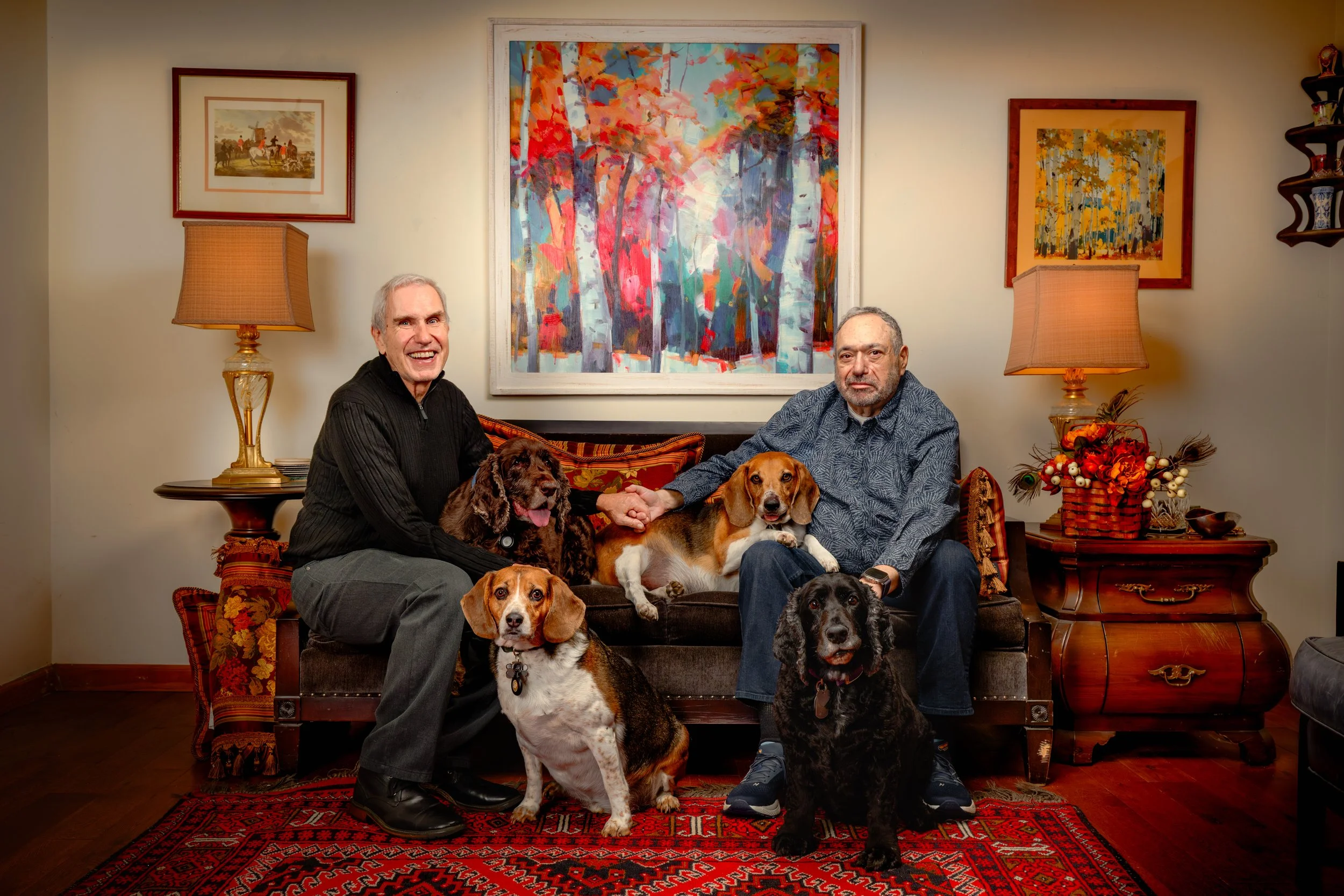 Two older men sitting together holding hands on a vintage sofa surrounded by five dogs, in a warmly decorated living room with colorful artwork and lamps.