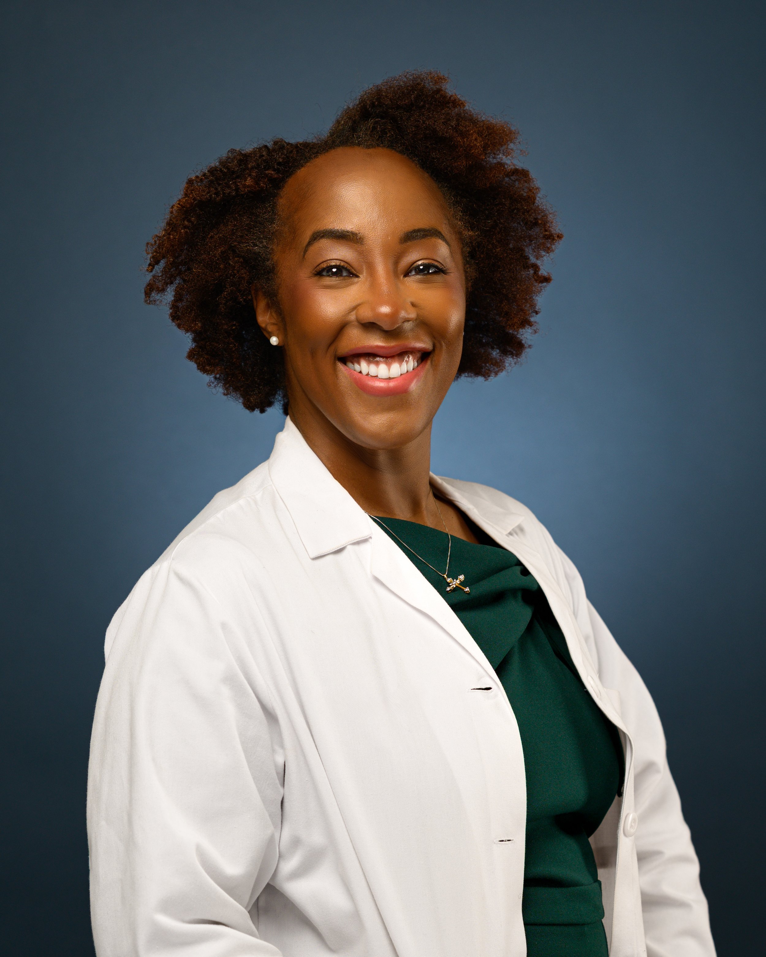 A headshot of a smiling woman with natural curly hair wearing a white lab coat over a green blouse, standing against a dark blue background.