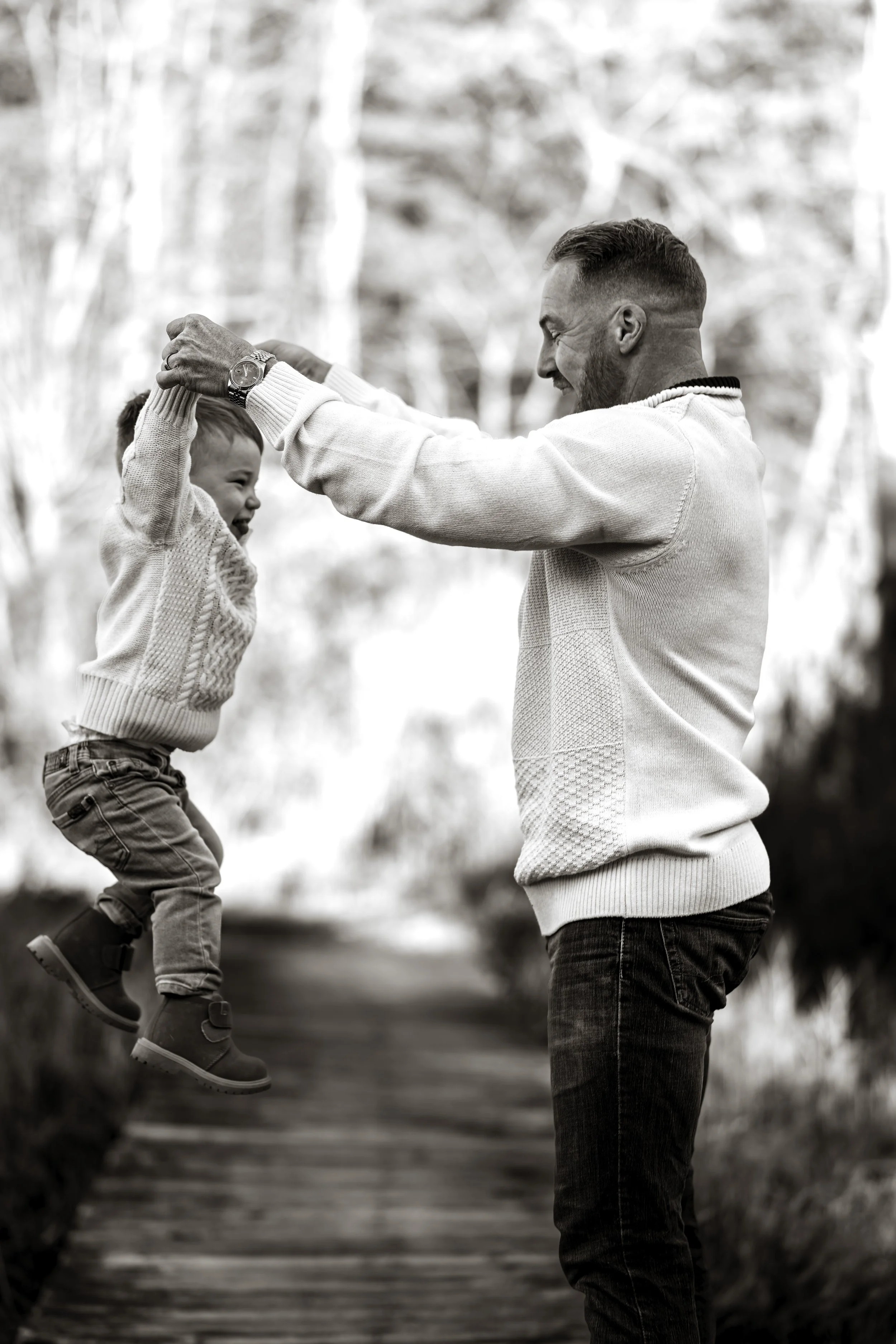 A man plays with a young child outdoors on a wooden walkway, lifting the child into the air. Both are smiling and appear happy, with trees in the background, photographed in black and white.