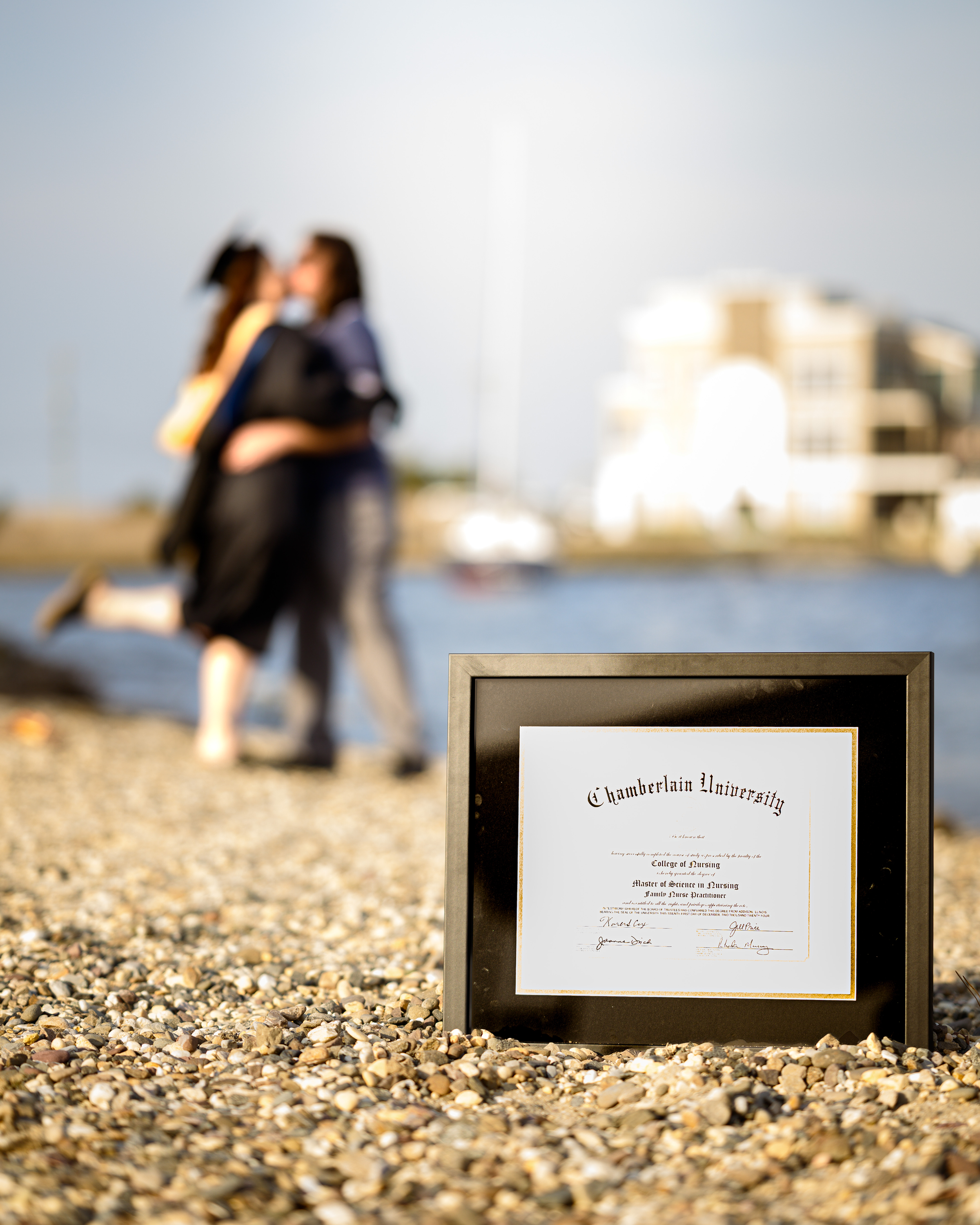 Framed diploma from Chamberlain University placed on a pebble beach with blurred graduates in caps and gowns walking near water and modern buildings in the background.