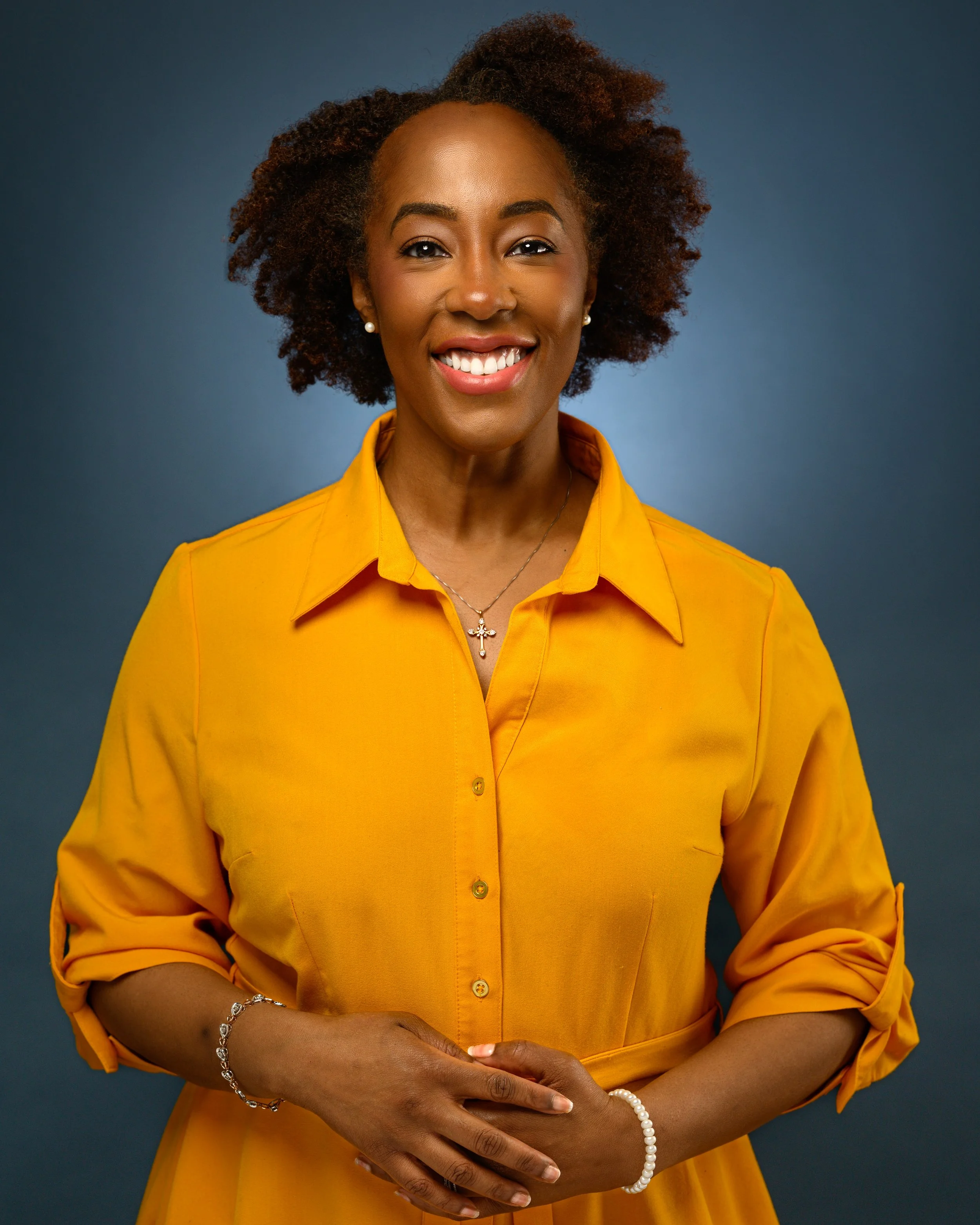 A headshot of awoman with curly hair and a bright smile wearing a yellow shirt, standing against a gray background.