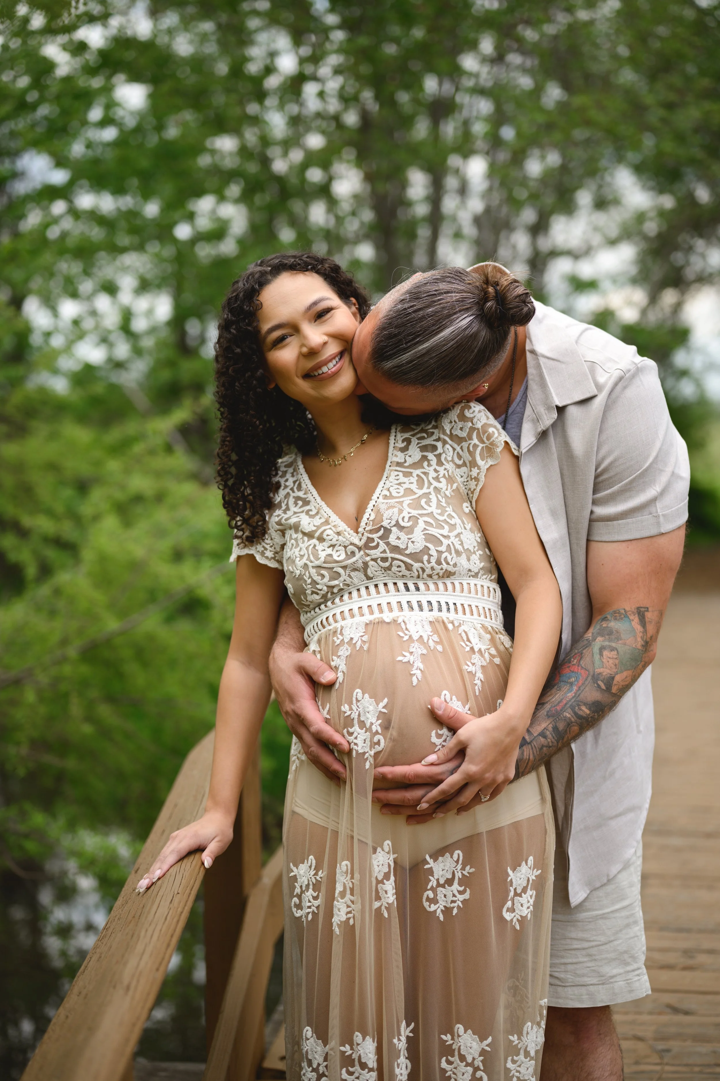 A couple standing on a wooden bridge in a park, the woman is pregnant and smiling, the man is kissing her cheek, surrounded by green trees.