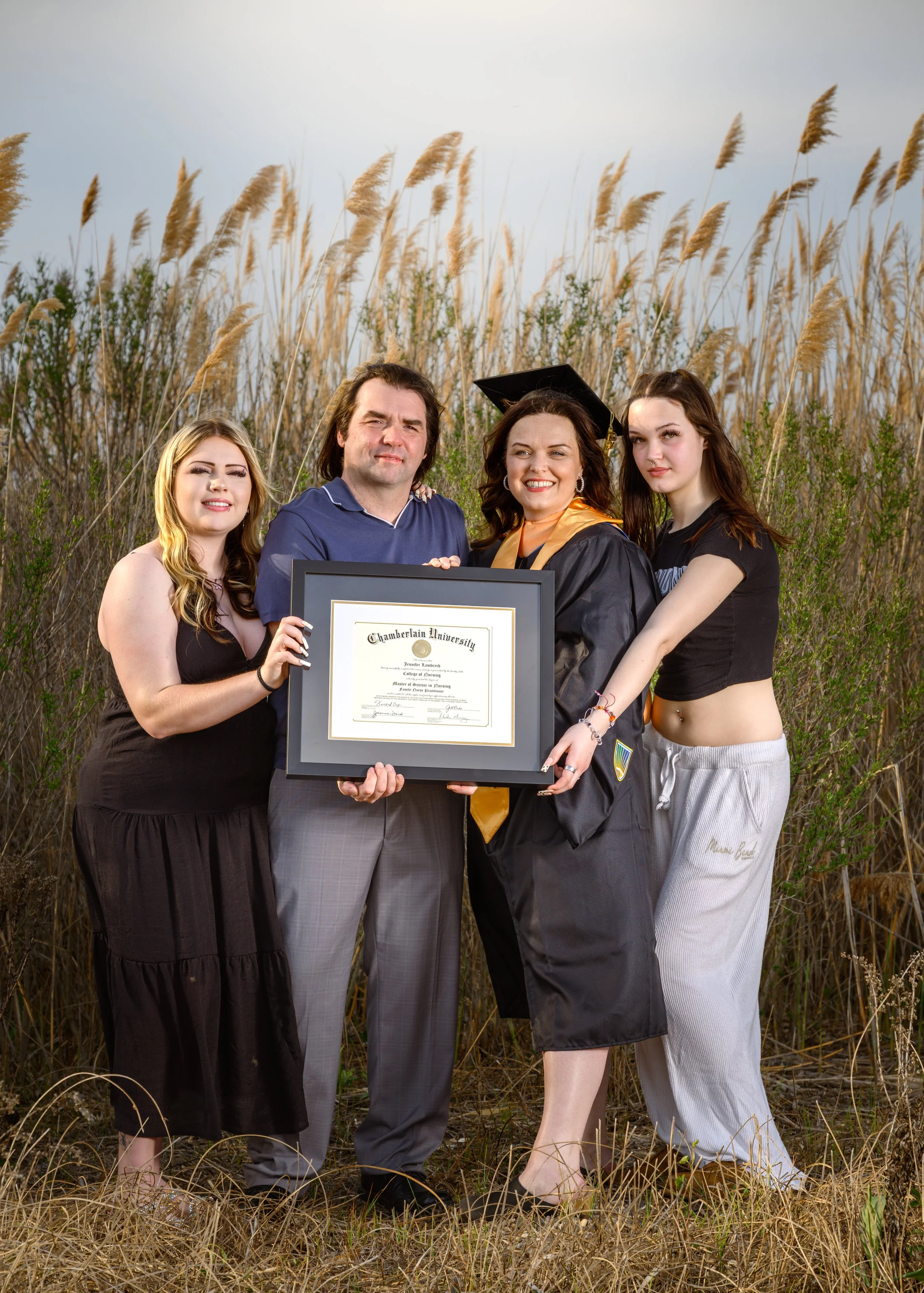 A woman in a cap and gown holding a framed diploma, standing with three other people in a field of tall grass.