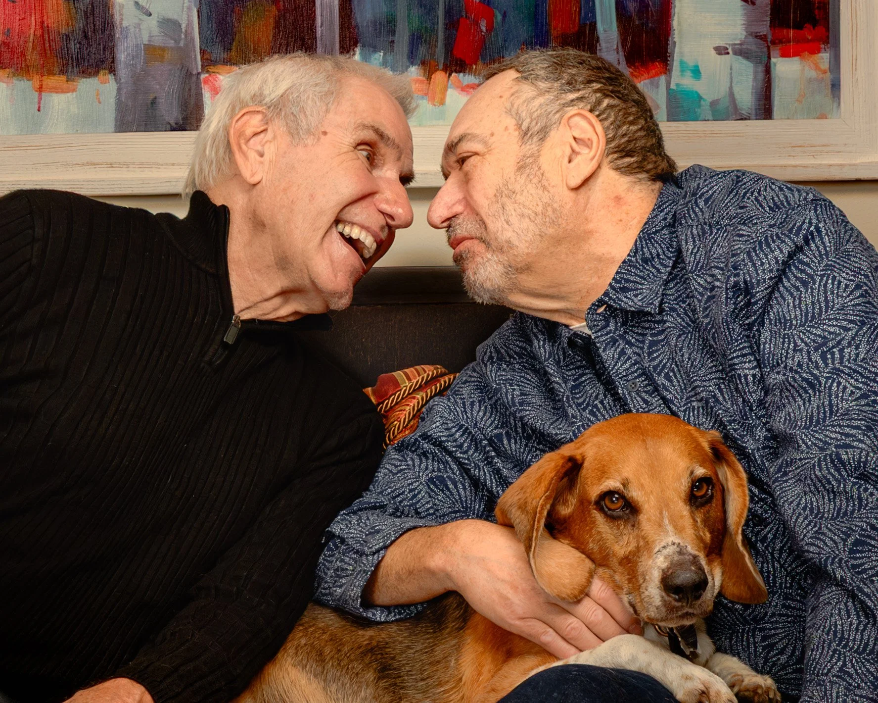 Two older men are close to each other, smiling and touching foreheads, with a brown dog lying between them, in a cozy indoor setting with colorful artwork on the wall in the background.