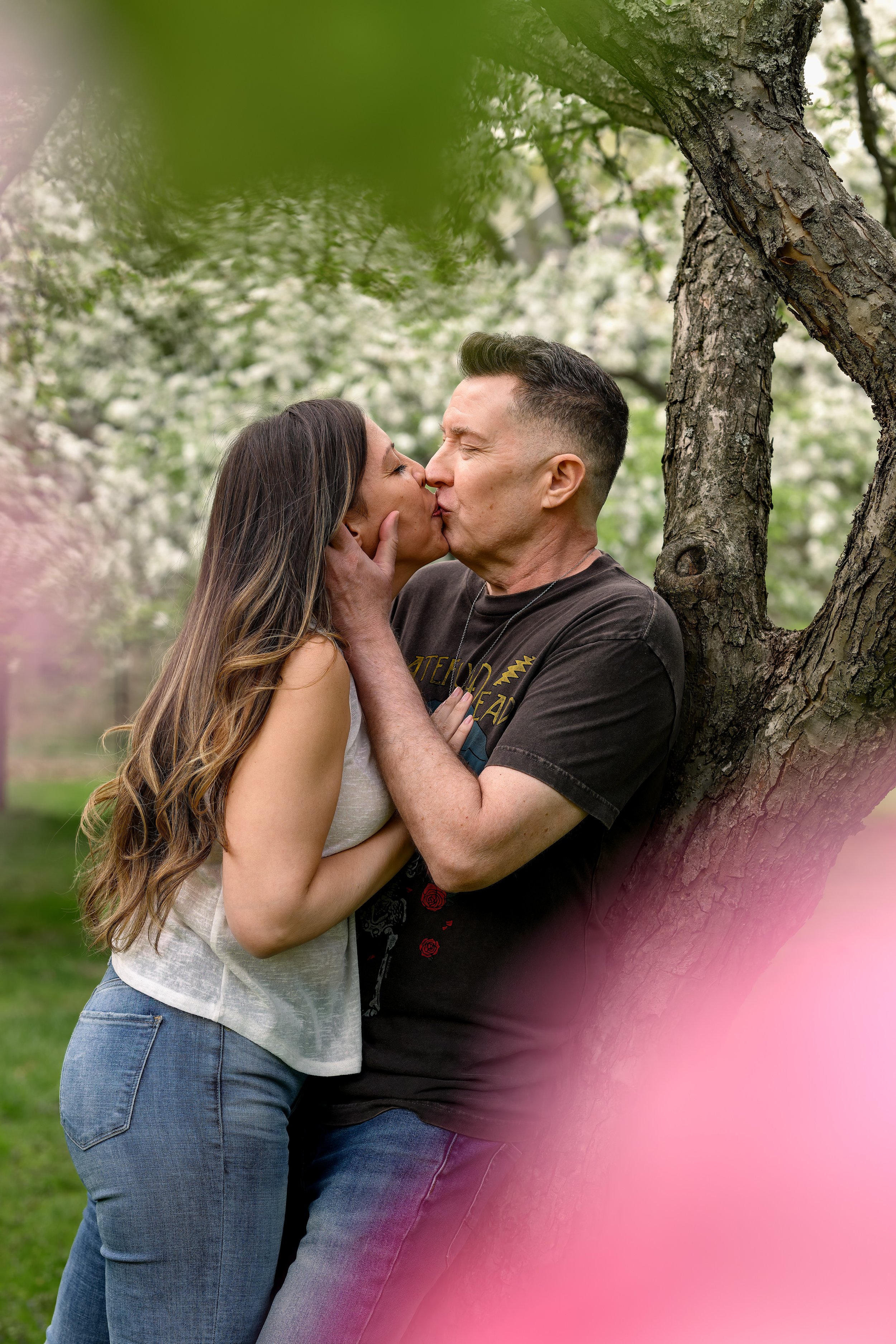 A couple sharing a kiss outdoors near a tree with blooming flowers, pink bloom in foreground.