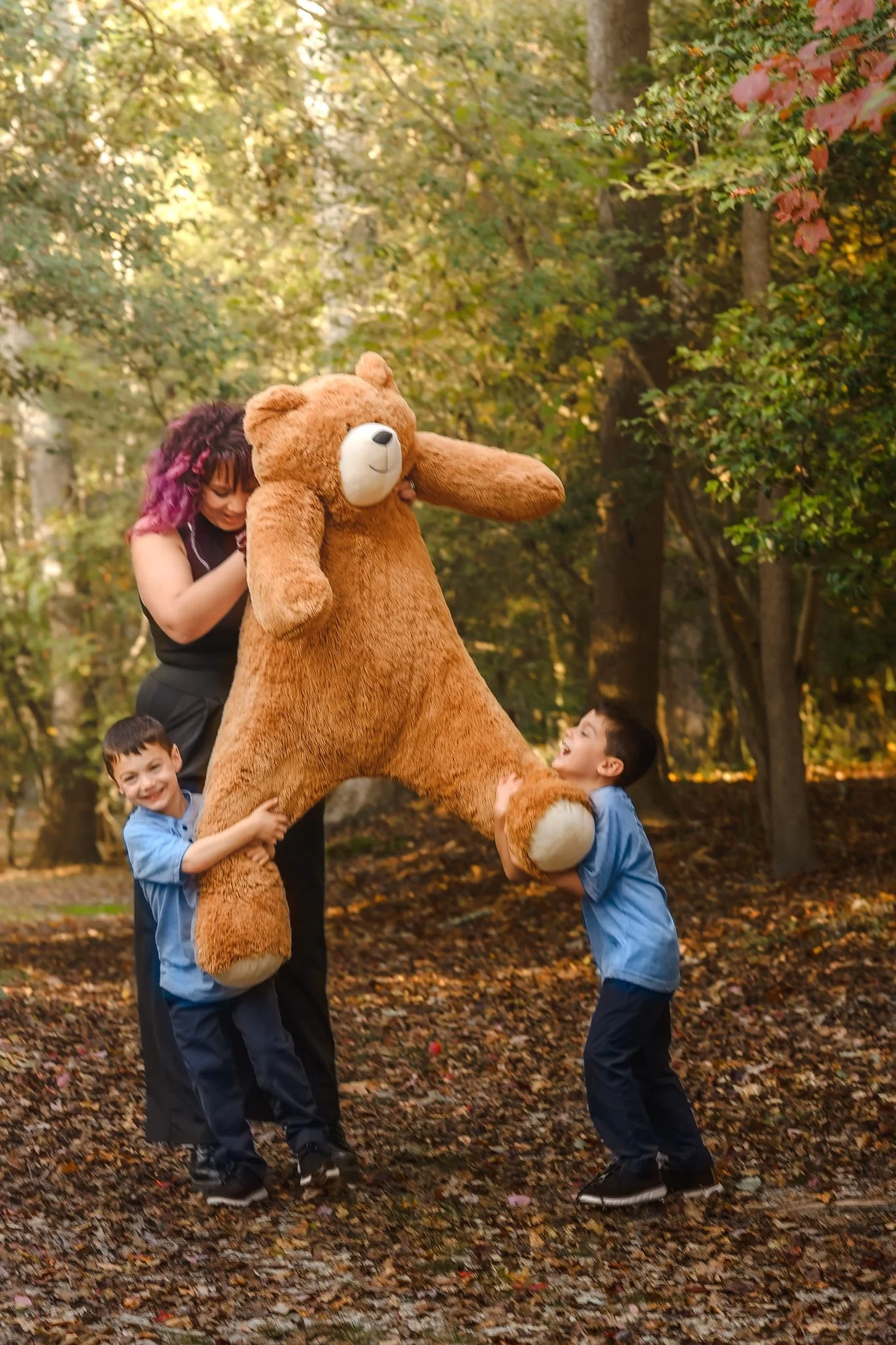 Two children and an adult playing with a large teddy bear in a wooded park during fall.