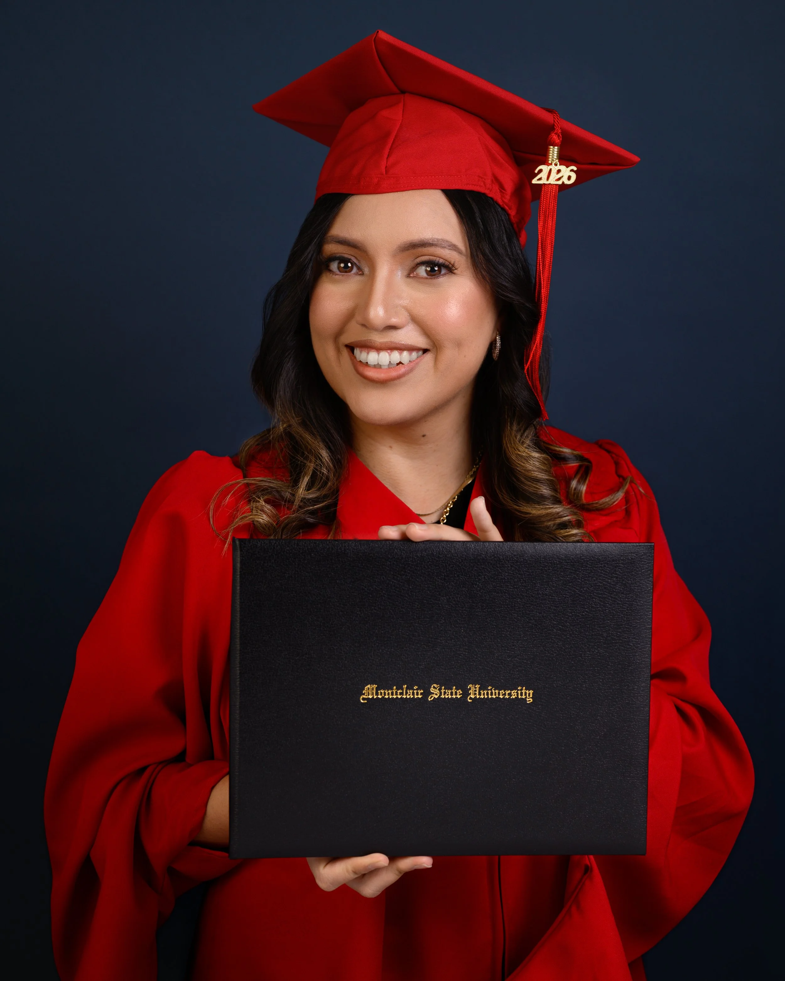 A woman in a red graduation cap and gown, holding a black diploma cover from Montclair State University, smiles at the camera against a dark background.