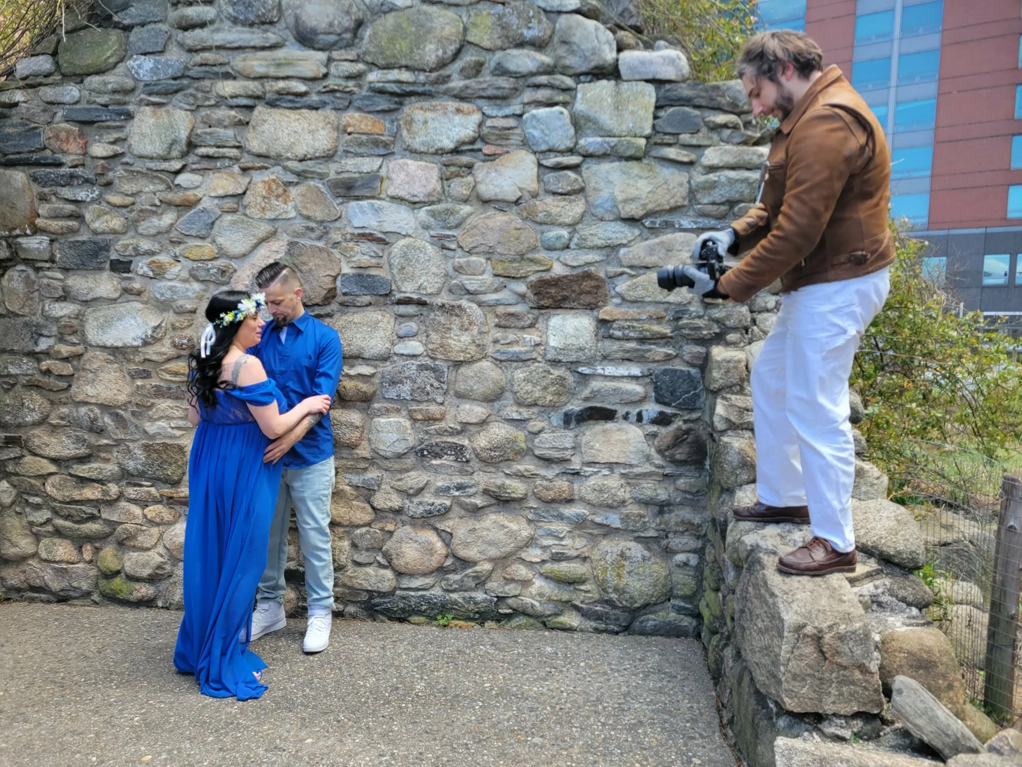 A couple, dressed in blue, standing close to each other against a stone wall, being photographed by a man with a camera on a rock ledge. The woman wears a long blue dress and a flower crown, and the man wears a blue shirt and light-colored pants.