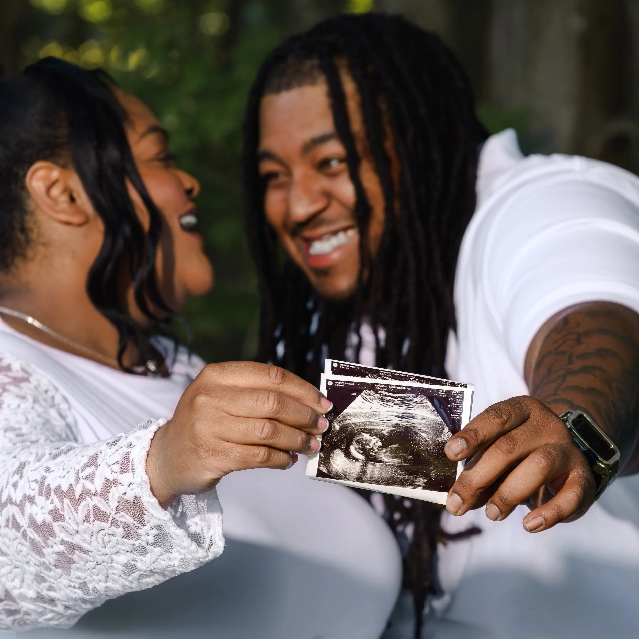 Close-up of a couple smiling and holding ultrasound photos outdoors, with a blurred background of greenery and water.