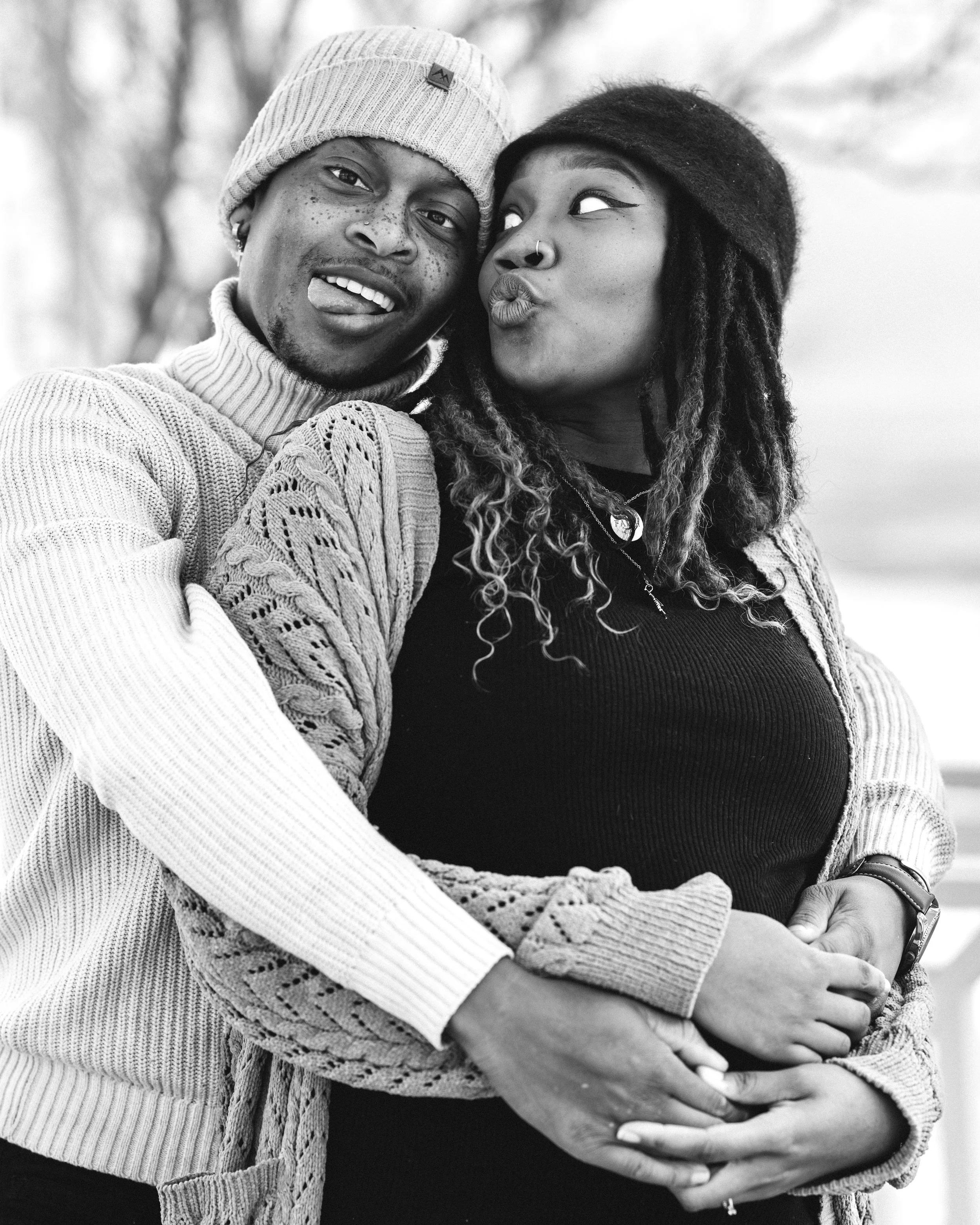 A black and white stylized photo of a married pregnant couple embracing each other and the mother's baby bump, making silly faces at each other and the camera