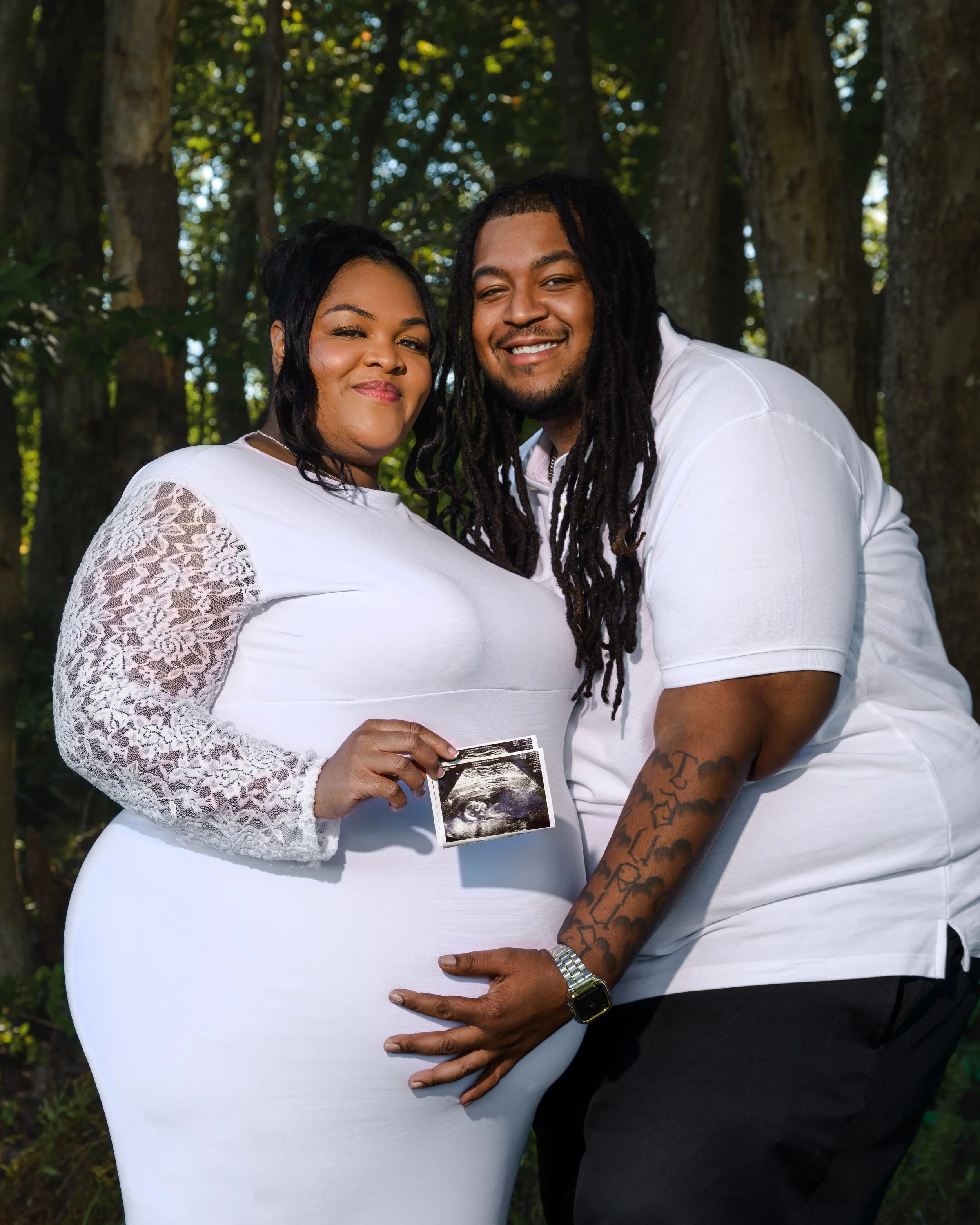 A couple standing outdoors in front of trees, smiling and holding an ultrasound photo, with the woman wearing a white dress with lace sleeves and the man wearing a white t-shirt.