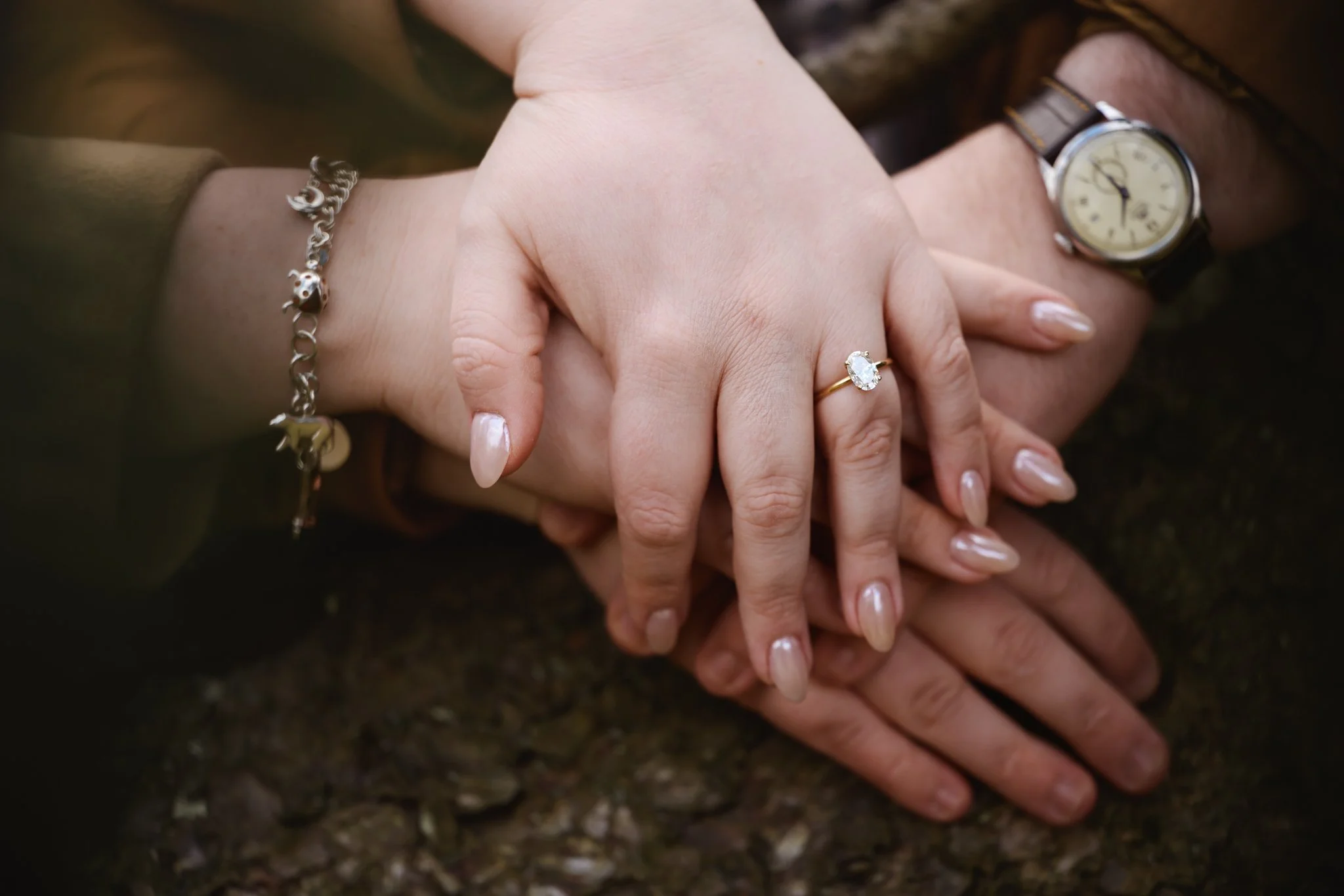 Close-up of an engaged couple holding hands, showing off the woman's diamond engagement ring. Their hands are resting on a warm-colored fir tree