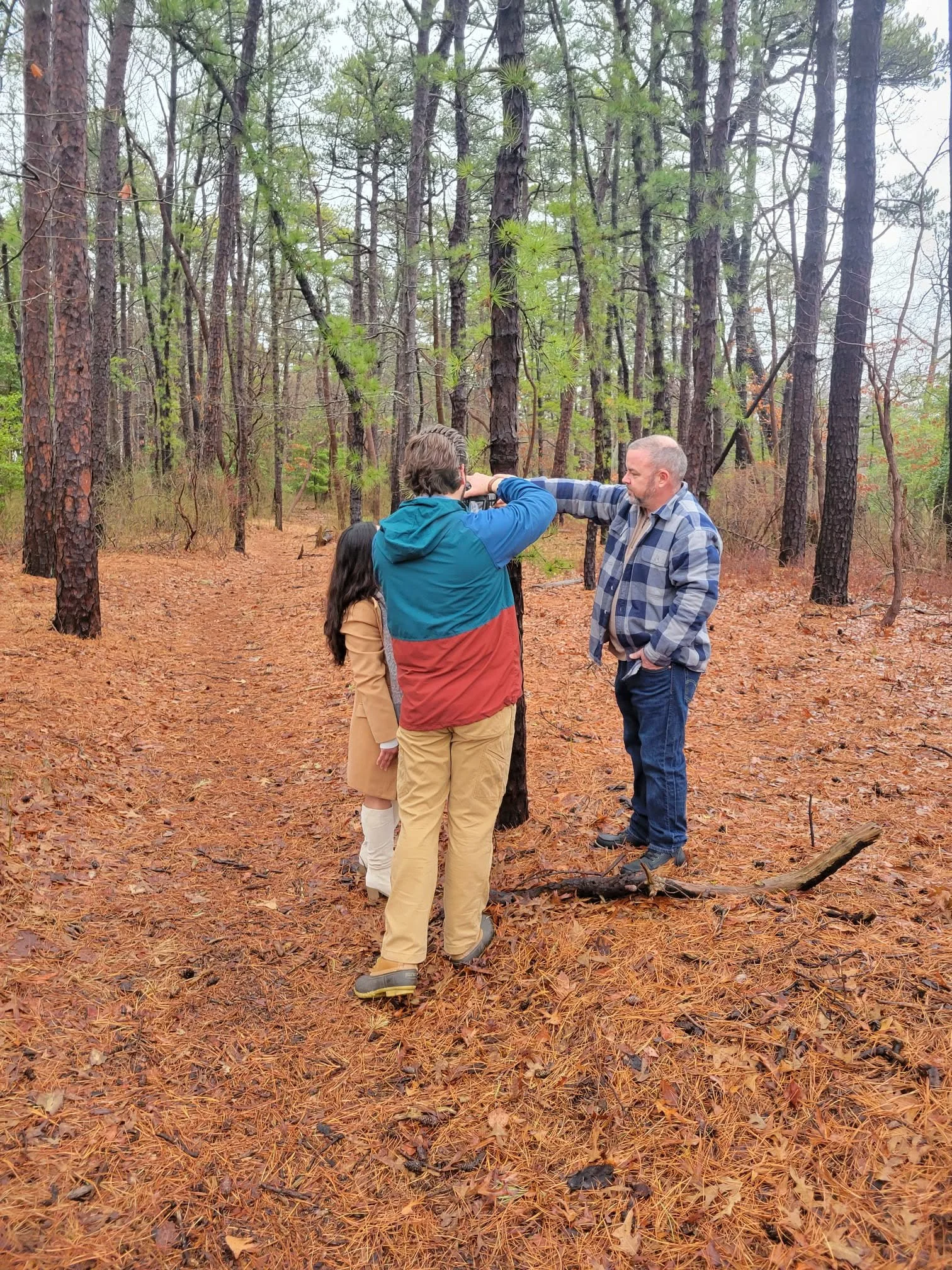 Three people standing in a forest, with one holding a camera to take a picture of the other two who are standing close together, surrounded by tall trees and orange pine needles on the ground.