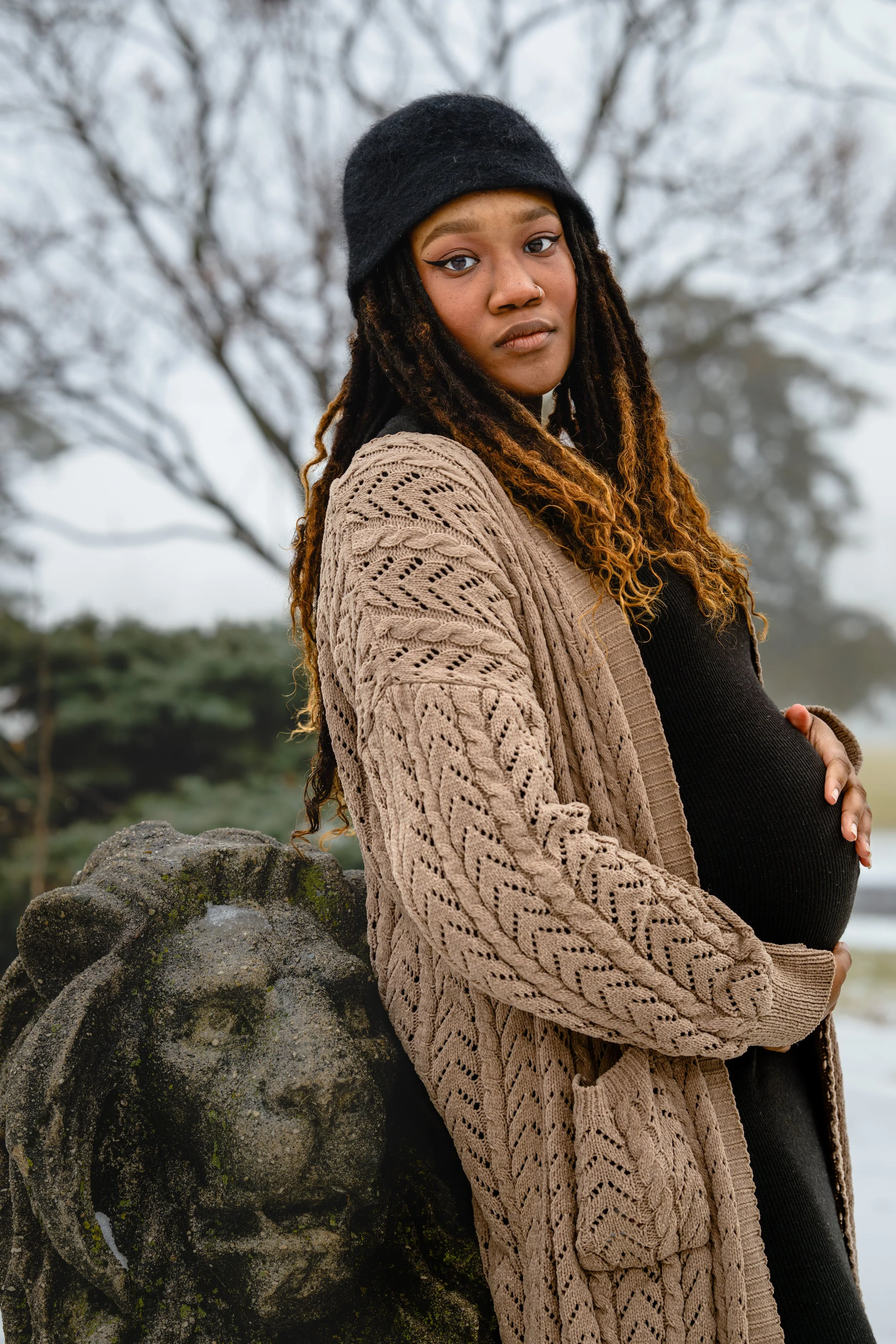 A pregnant woman in a cable-knit cardigan and black hat, holding her baby bump and leaning against a lion statue in the snow outside while looking directly into the camera