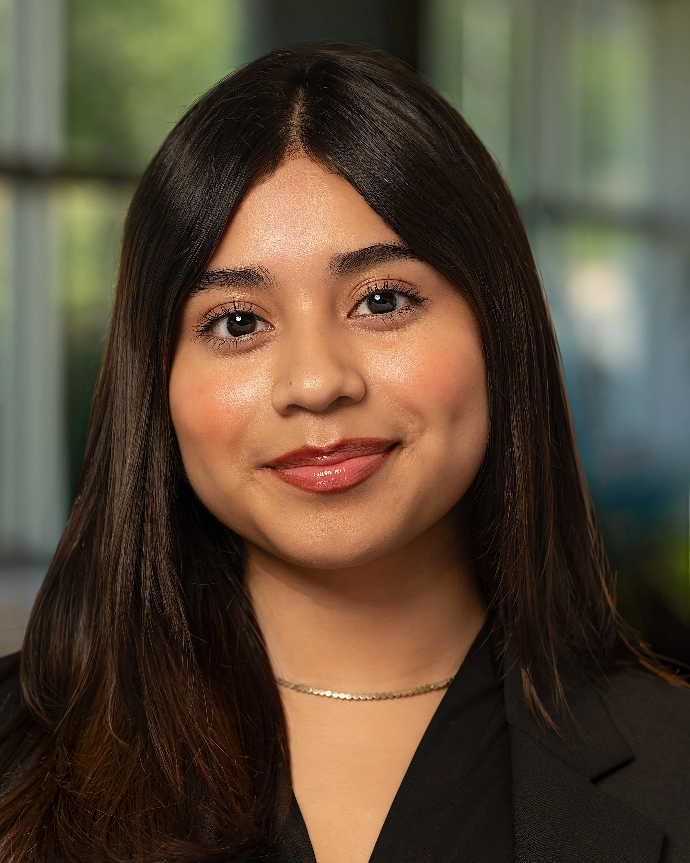 Close-up headshot of a smiling young woman with long dark hair and a black blazer, standing indoors with a blurred window background.