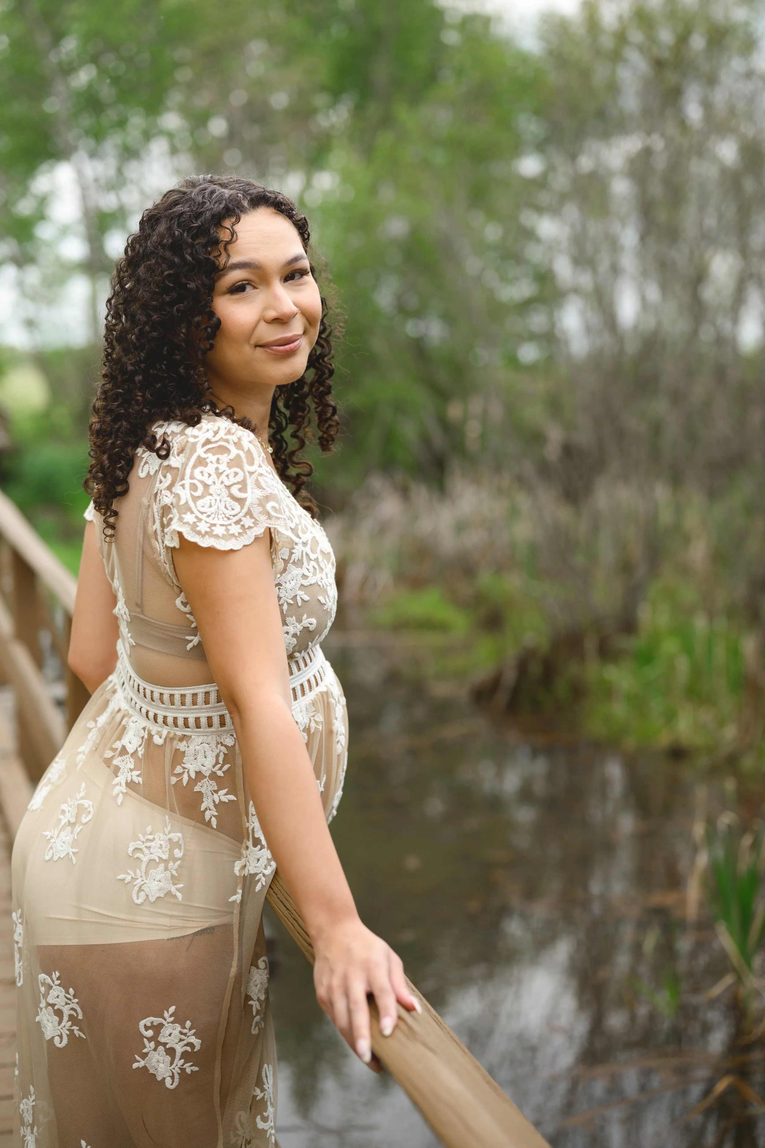 A pregnant young woman with curly hair wearing a beige lace dress standing on a wooden bridge near a small body of water in a green outdoor setting.