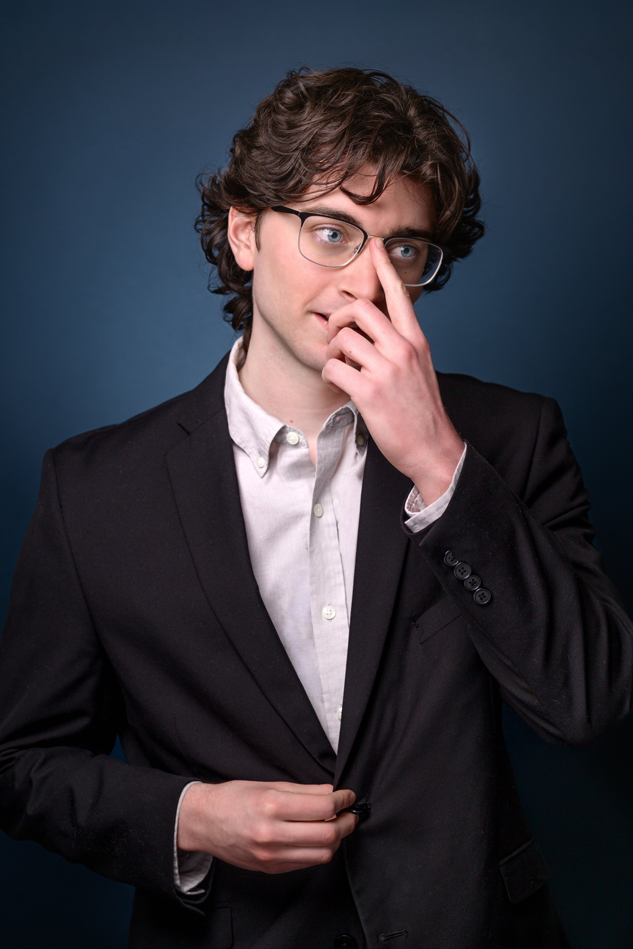 A man with curly brown hair and glasses, dressed in a black blazer and white shirt, adjusting his blazer with one hand while touching his nose with the other, standing against a dark blue background.