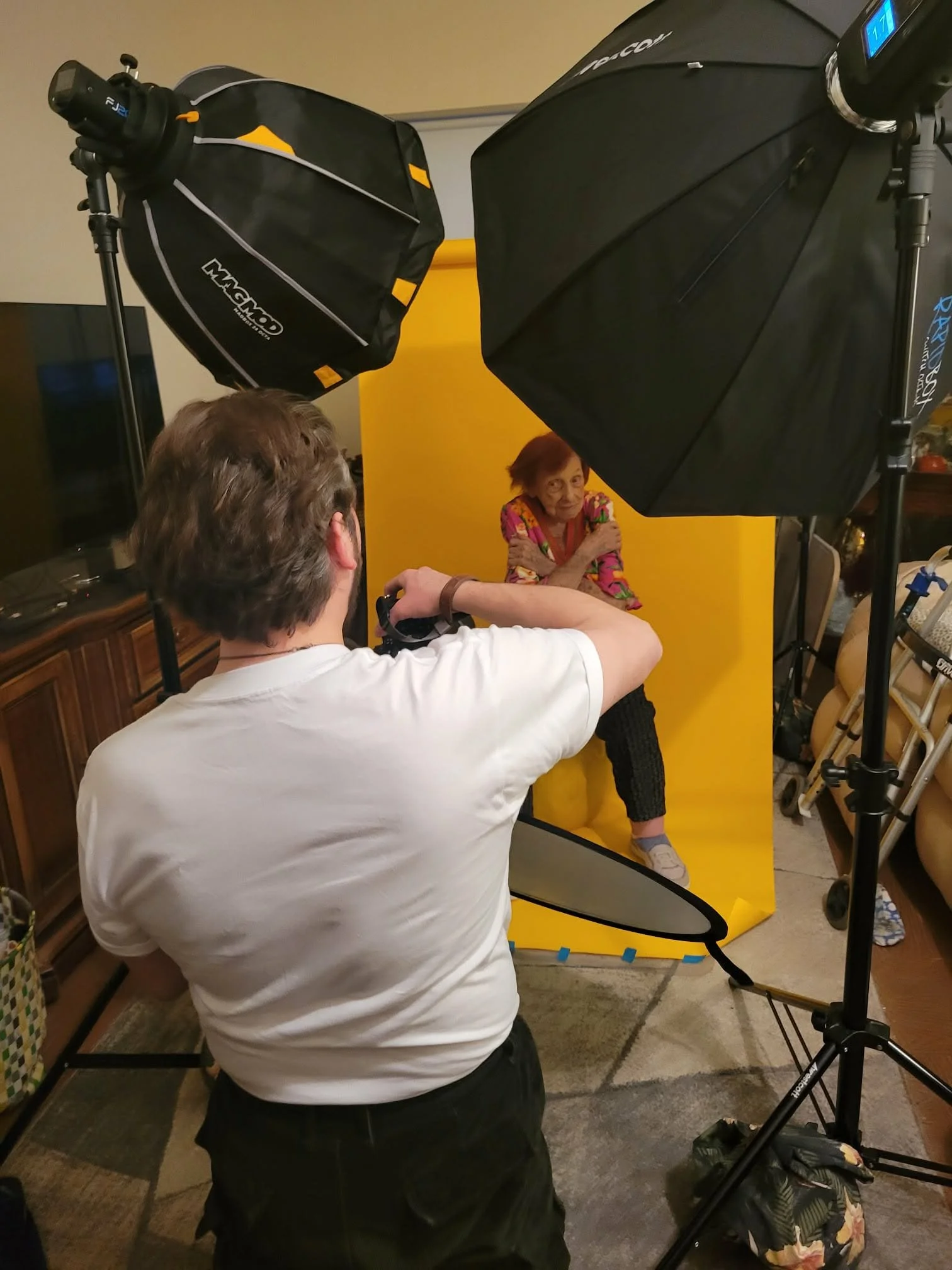 Photographer taking a picture of an elderly woman seated against a yellow background in a photo studio with lighting equipment.
