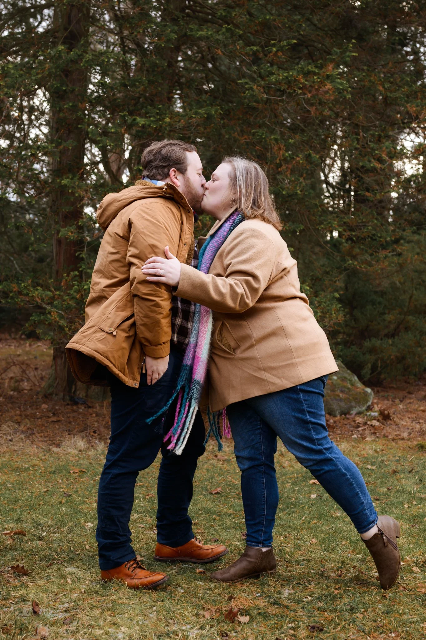 A couple wearing brown jackets sharing a kiss outdoors in a wooded area with green and brown foliage.