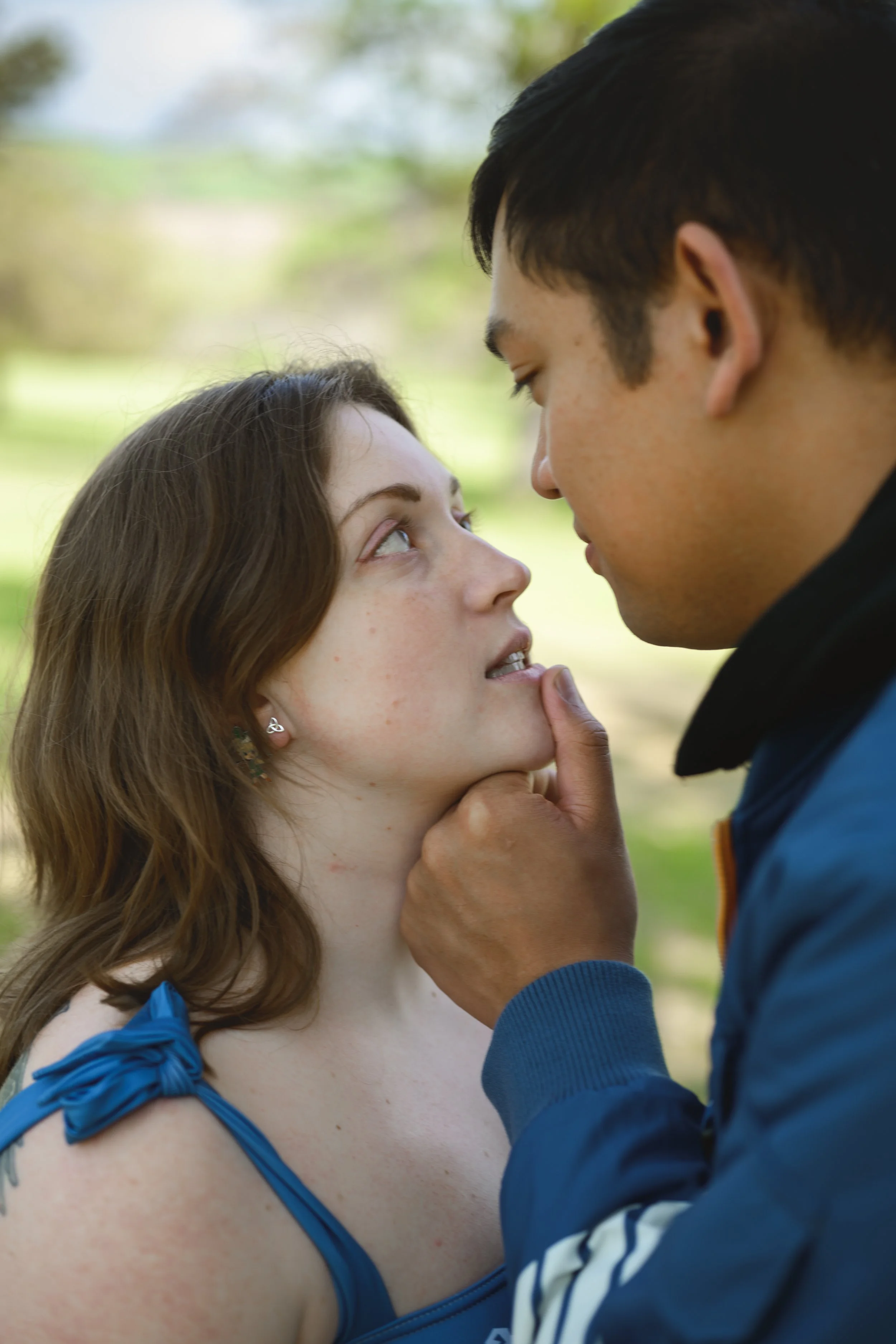 A young woman and a man are close together, about to kiss in a park with blurred green trees in the background.