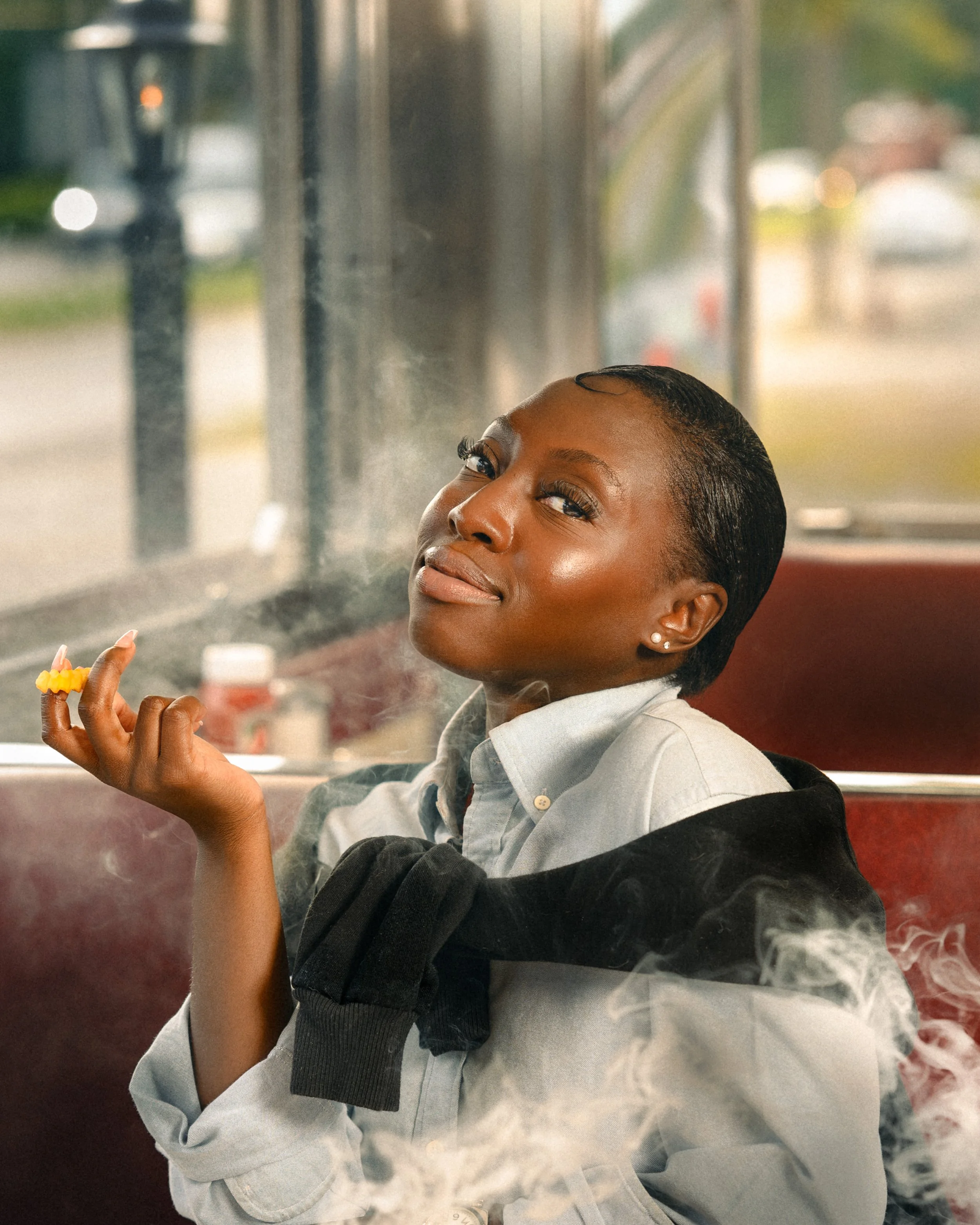 A woman with short hair and earrings at a diner, holding a French fry while smoke billows around her, as if coming from the French fry itself