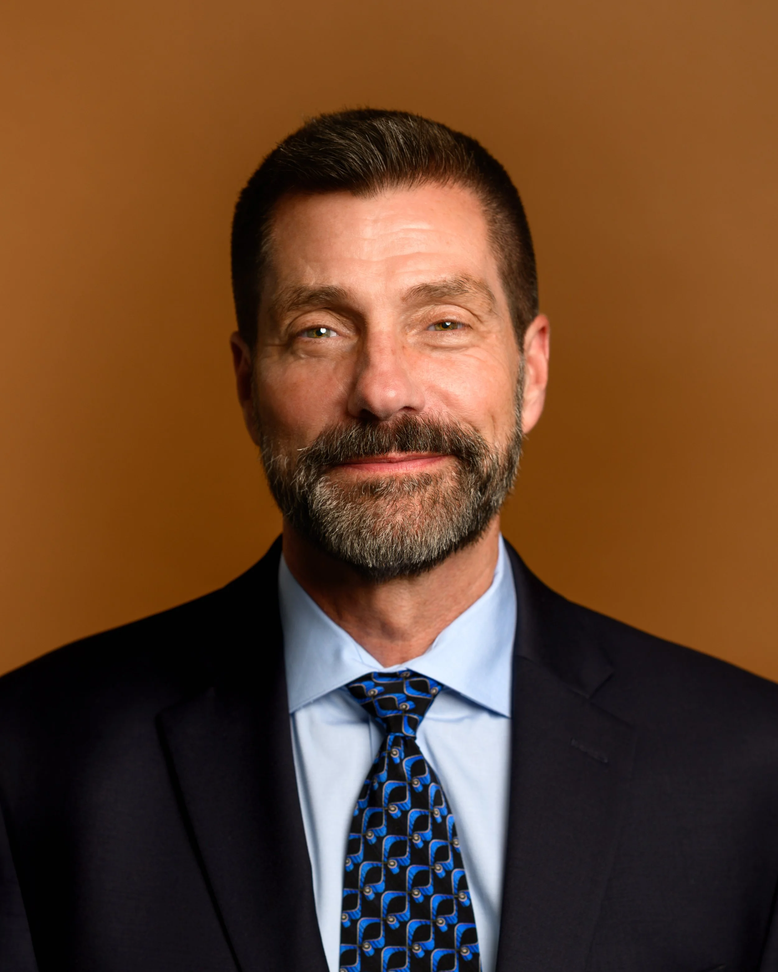 Portrait of a middle-aged man with a beard, wearing a black suit, light blue shirt, and a patterned blue tie, standing against a brown background.