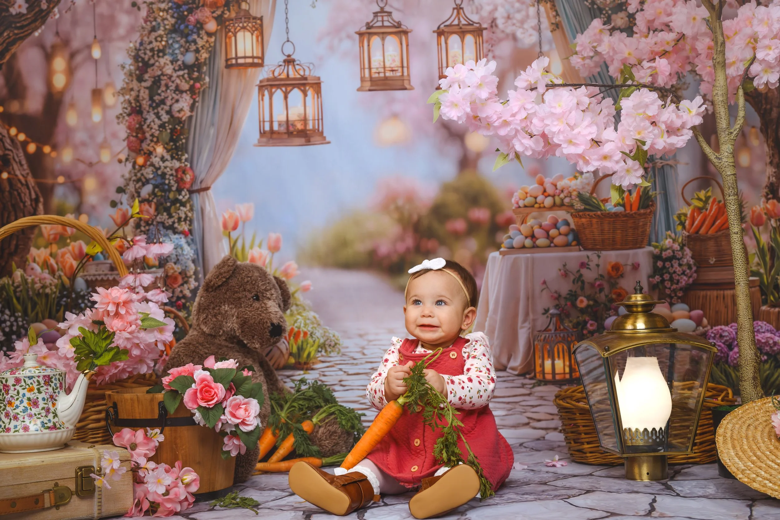 A baby girl sitting outdoors on a cobblestone path decorated with pink cherry blossoms, flowers, and lanterns. She is holding a large carrot and surrounded by a teddy bear, baskets, teapot, and floral arrangements, creating a whimsical garden scene.