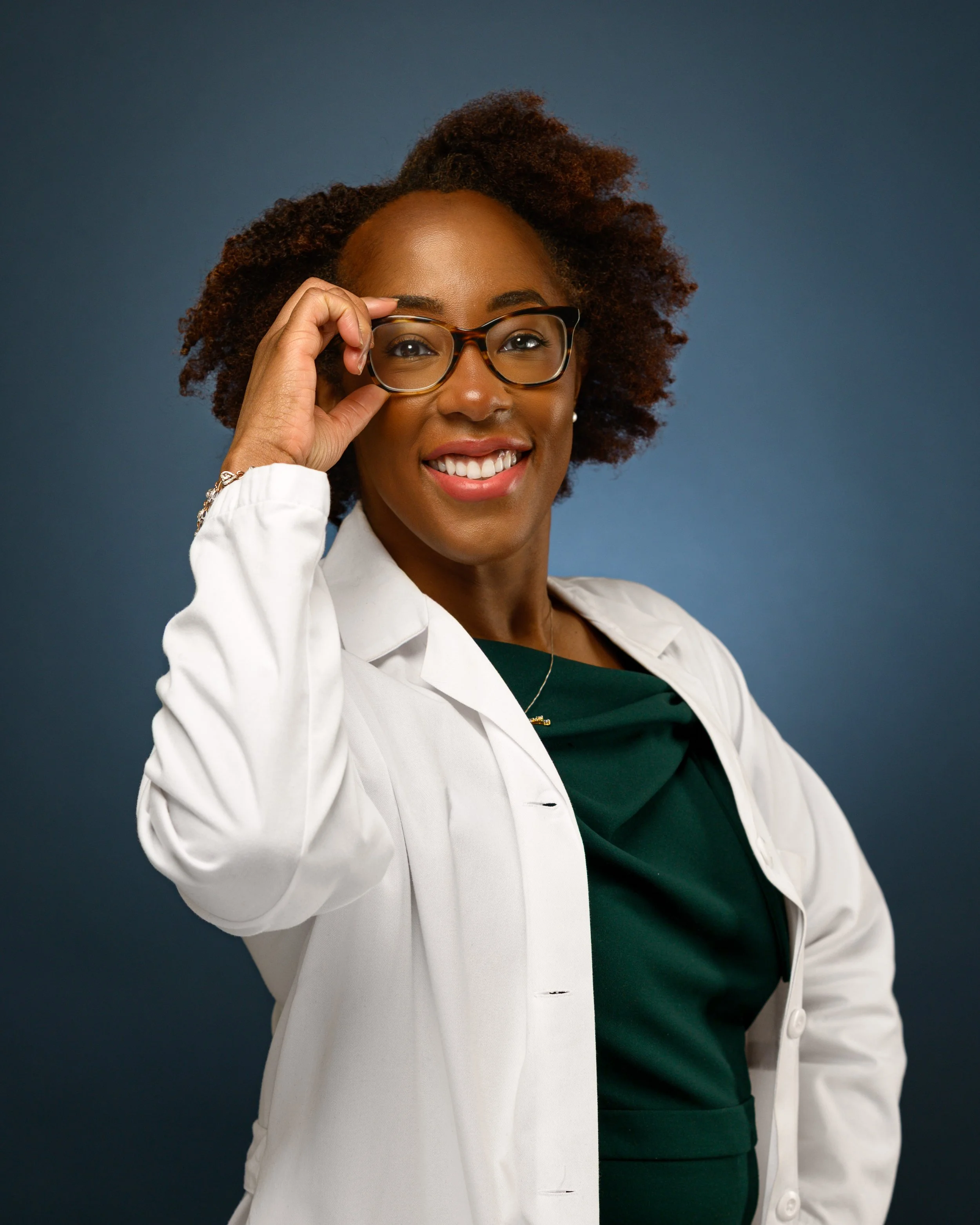 Headshot of a smiling woman with natural curly hair, wearing glasses, a white doctor's coat, and a dark green top, standing against a dark blue background.