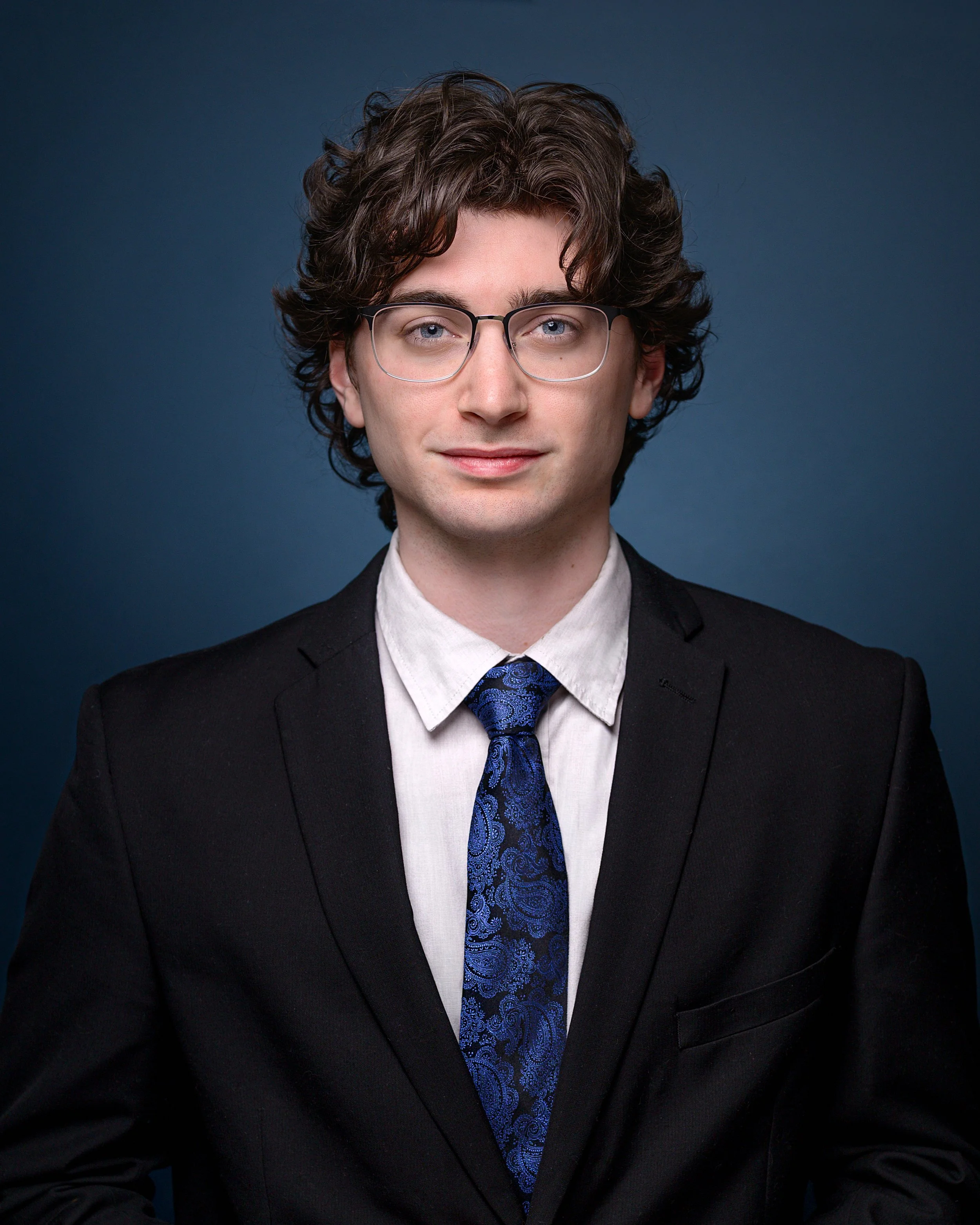 Portrait of a young man with glasses, wavy brown hair, dressed in a black suit, white shirt, and blue patterned tie, against a dark blue background.