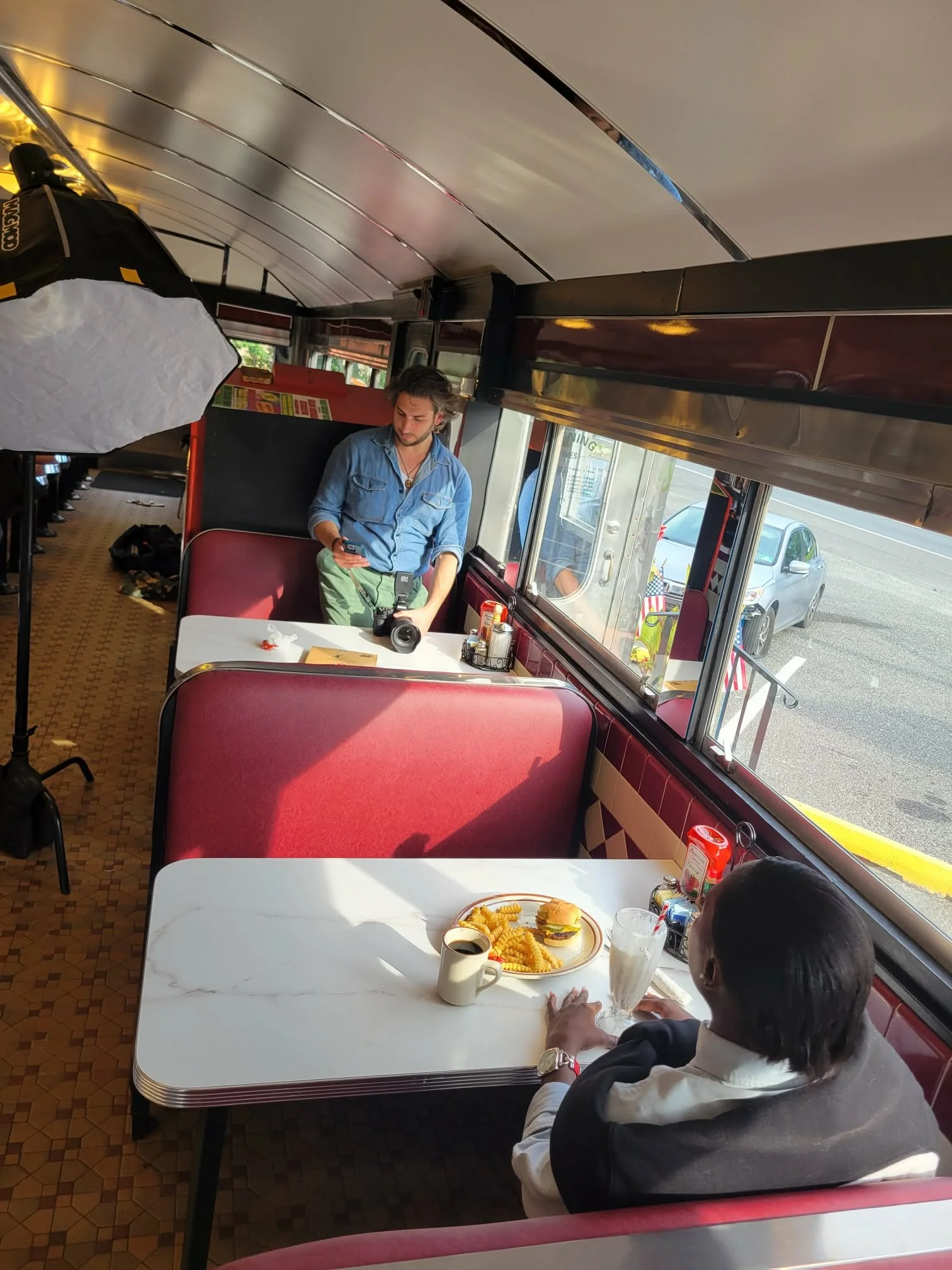 Inside a retro-style diner with red cushioned booths and white tables. A woman is sitting at a table with a frosted mug of coffee, a glass of water, and a tray with a burger, fries, and a cup of coffee. A young man is standing near the window, looking at his phone.