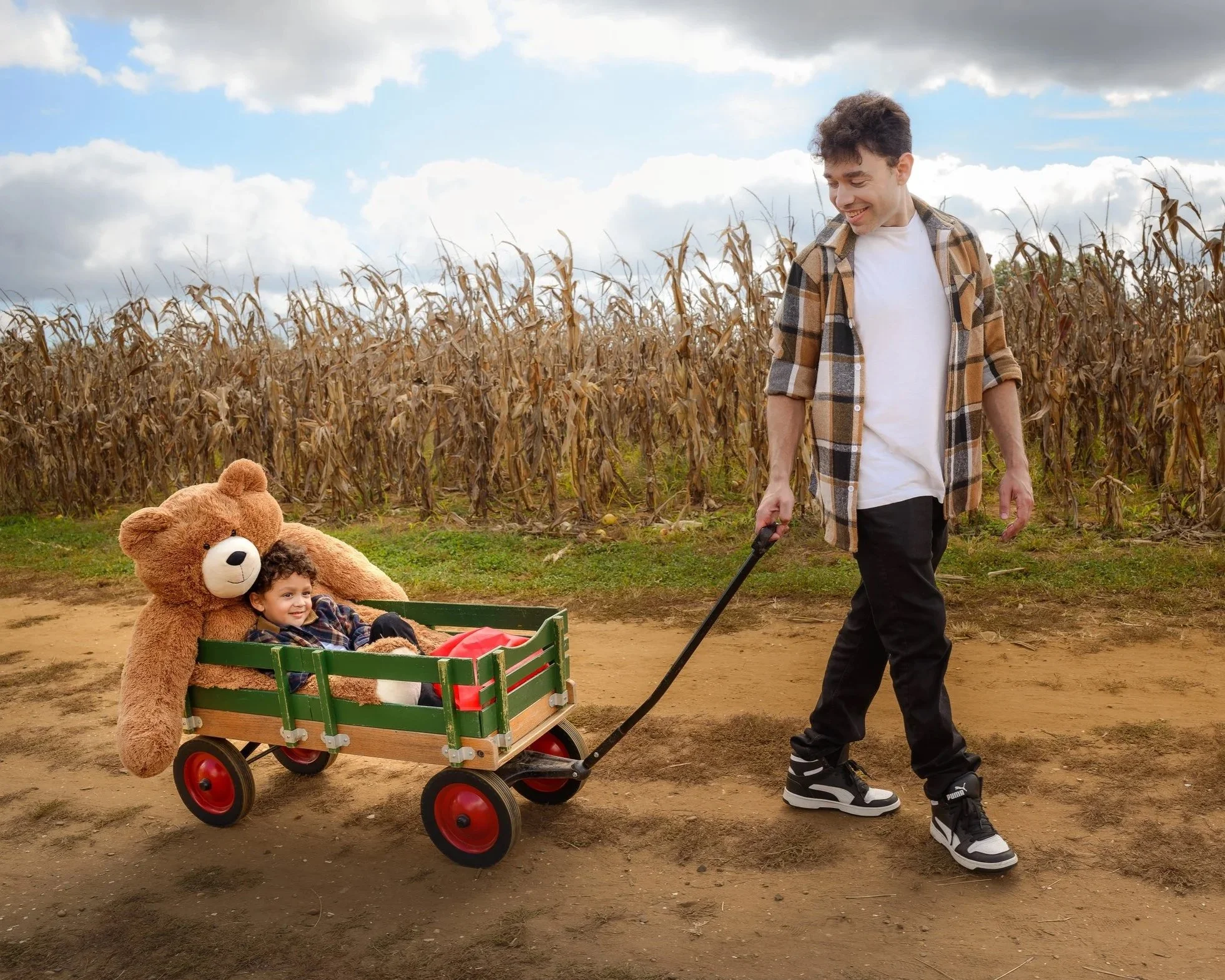 A smiling man is pulling a small wagon with a young child and a large teddy bear through a rural area with a cornfield in the background.