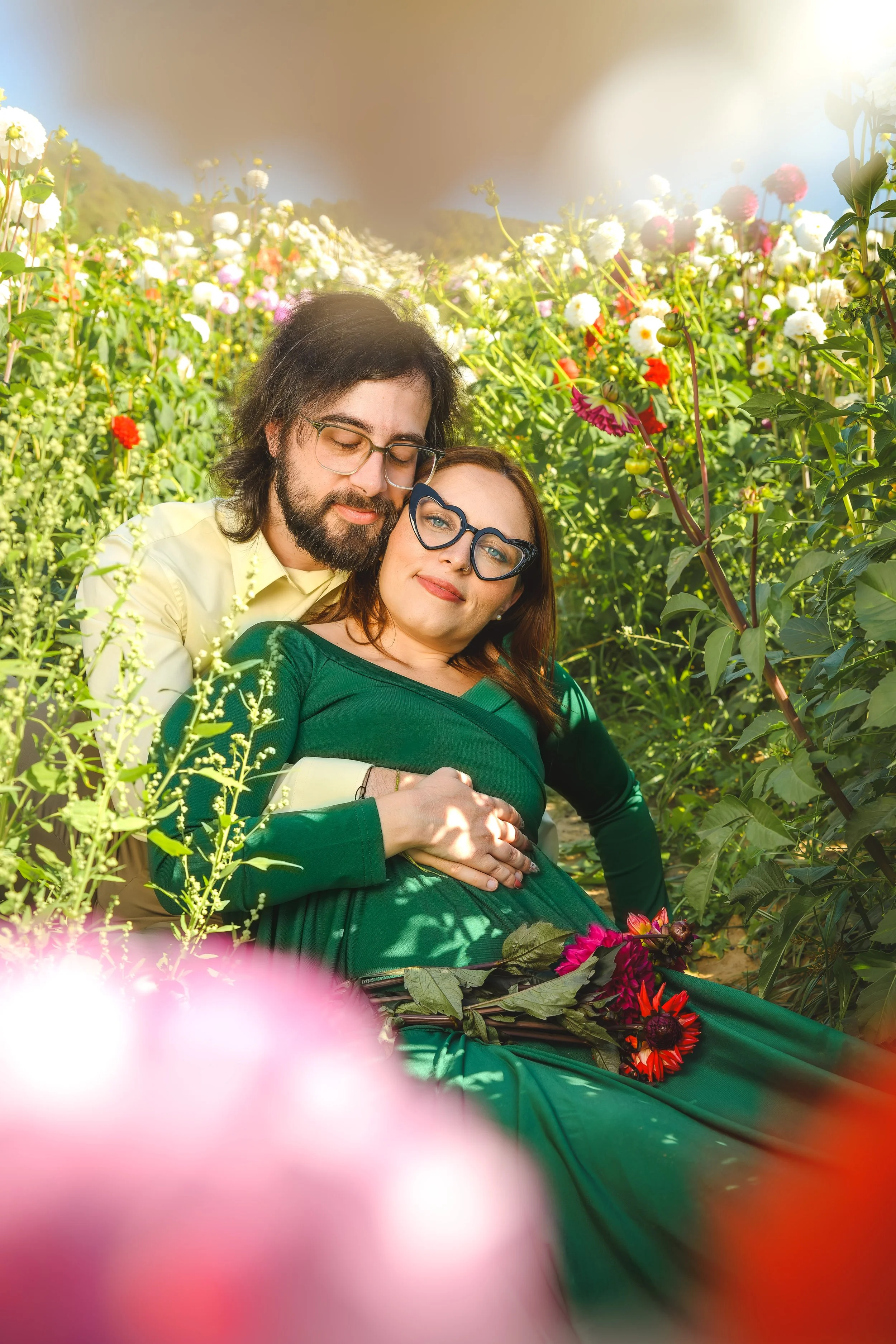 A pregnant woman and a man are sitting among colorful flowers in a garden, with the woman resting her hand on her belly and both smiling peacefully.