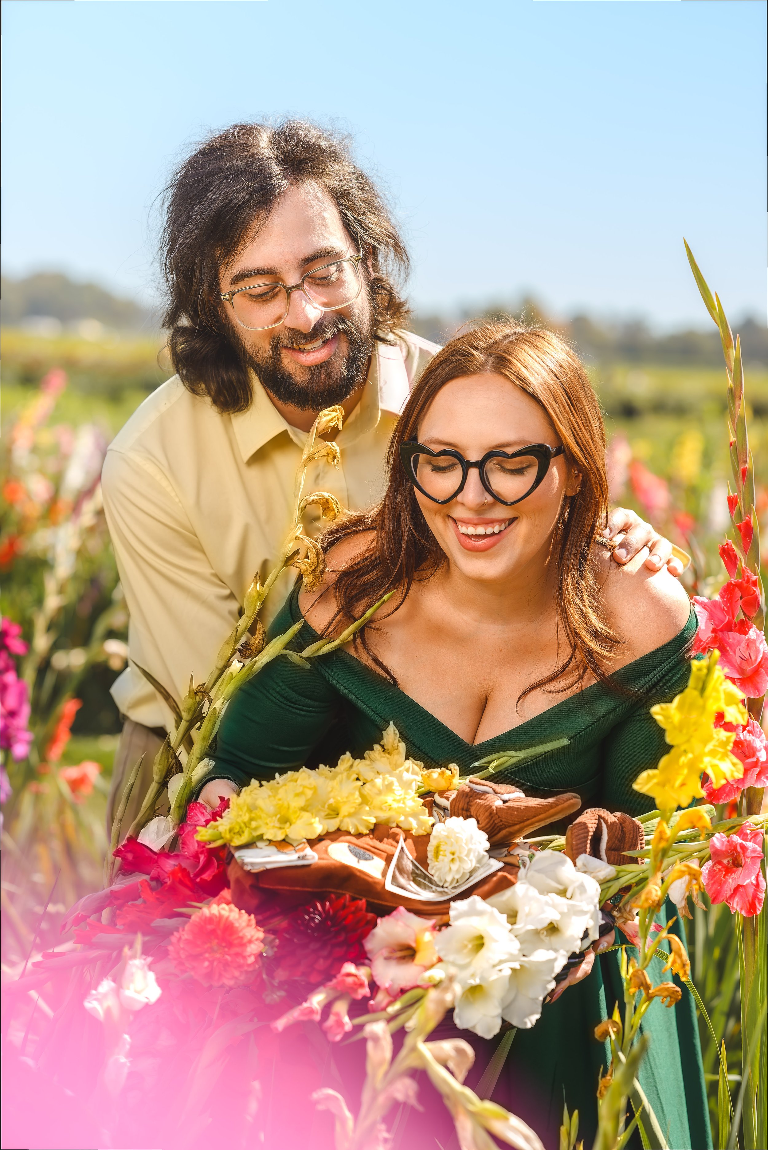 A man and woman in a flower garden, smiling and looking at flowers, with a basket of flowers and baked goods.