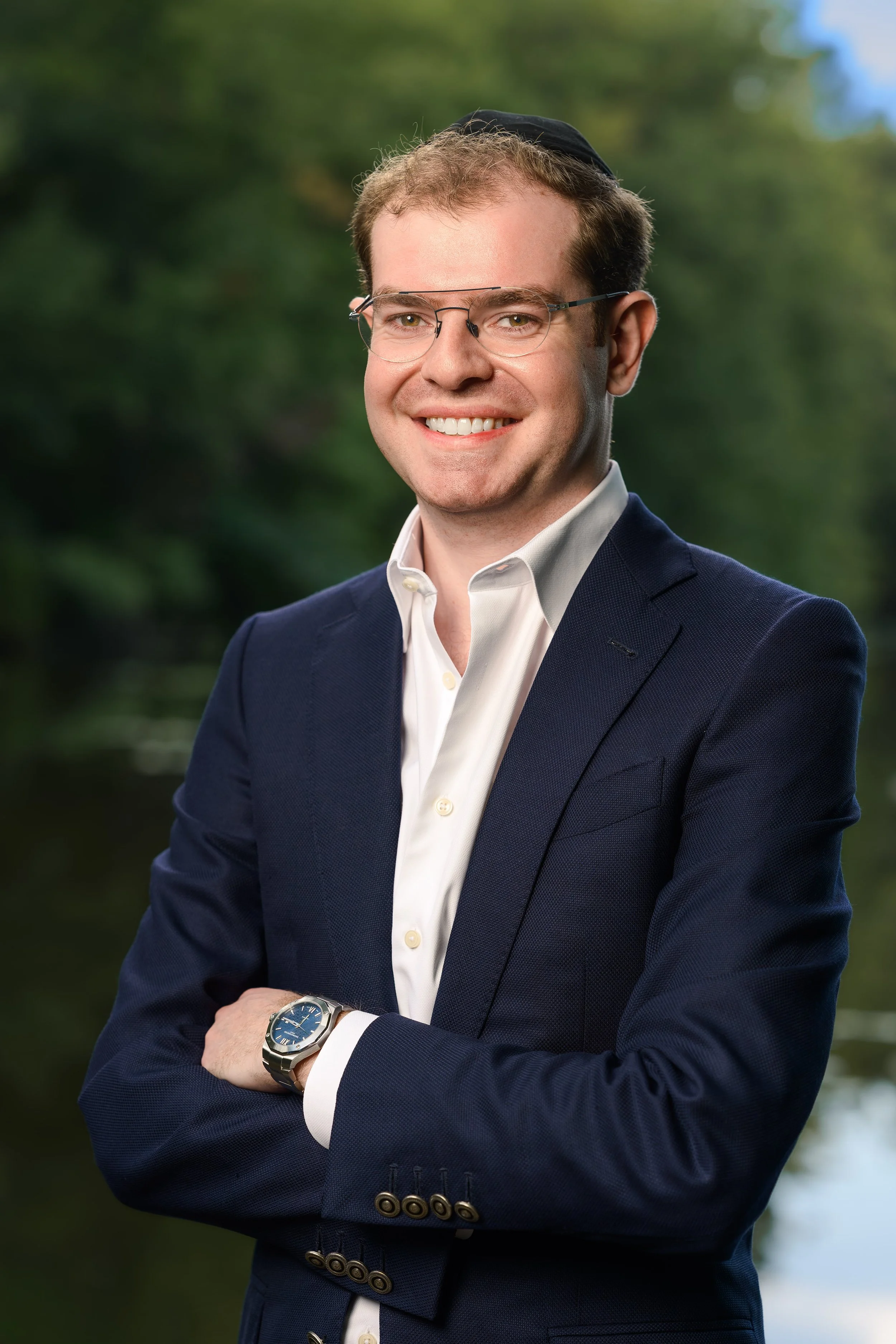 A headshot of a smiling man in a business suit and glasses standing outdoors with trees in the background.