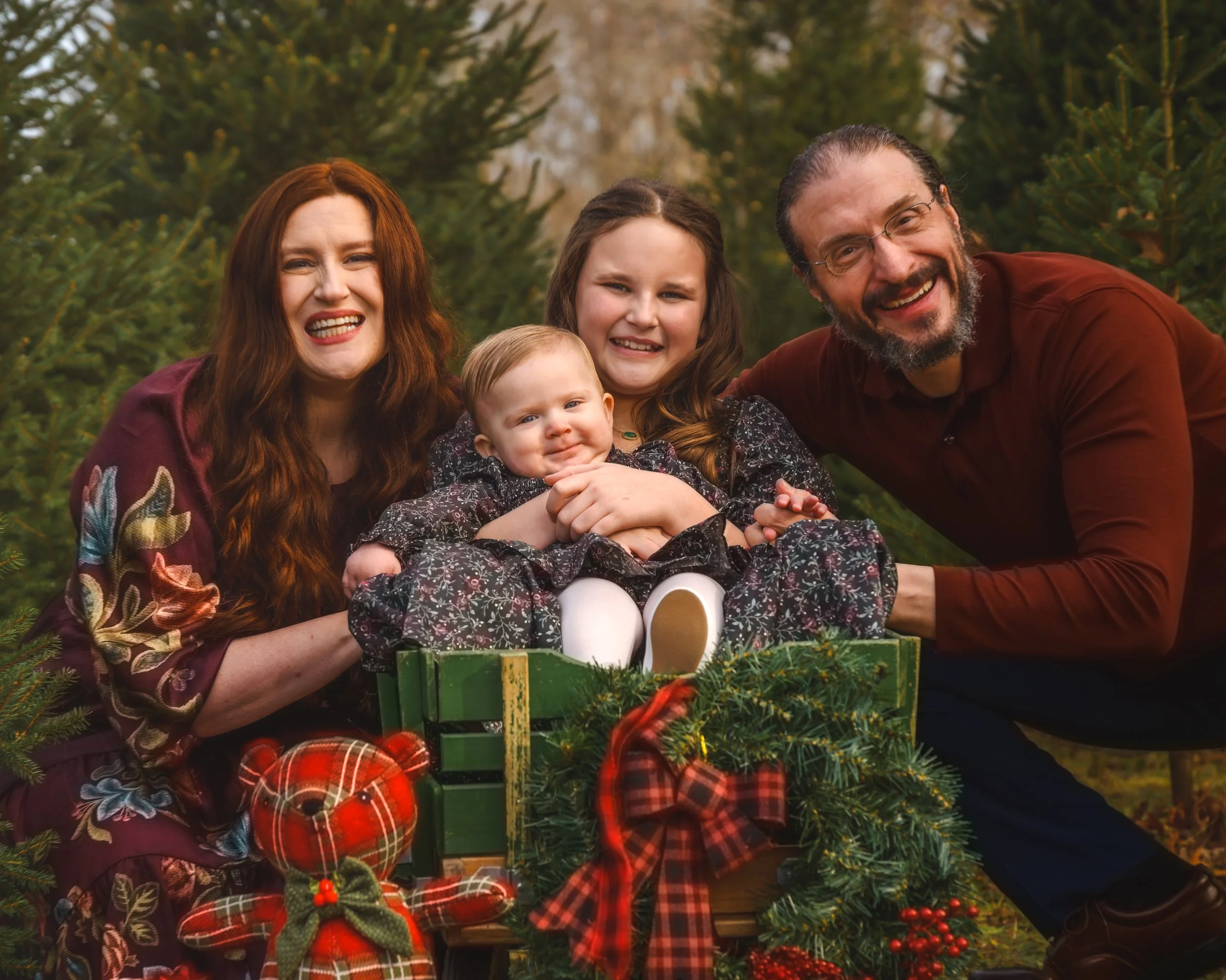 Family of four smiling outdoors next to a Christmas gift box decorated with greenery and red ribbons, surrounded by evergreen trees.