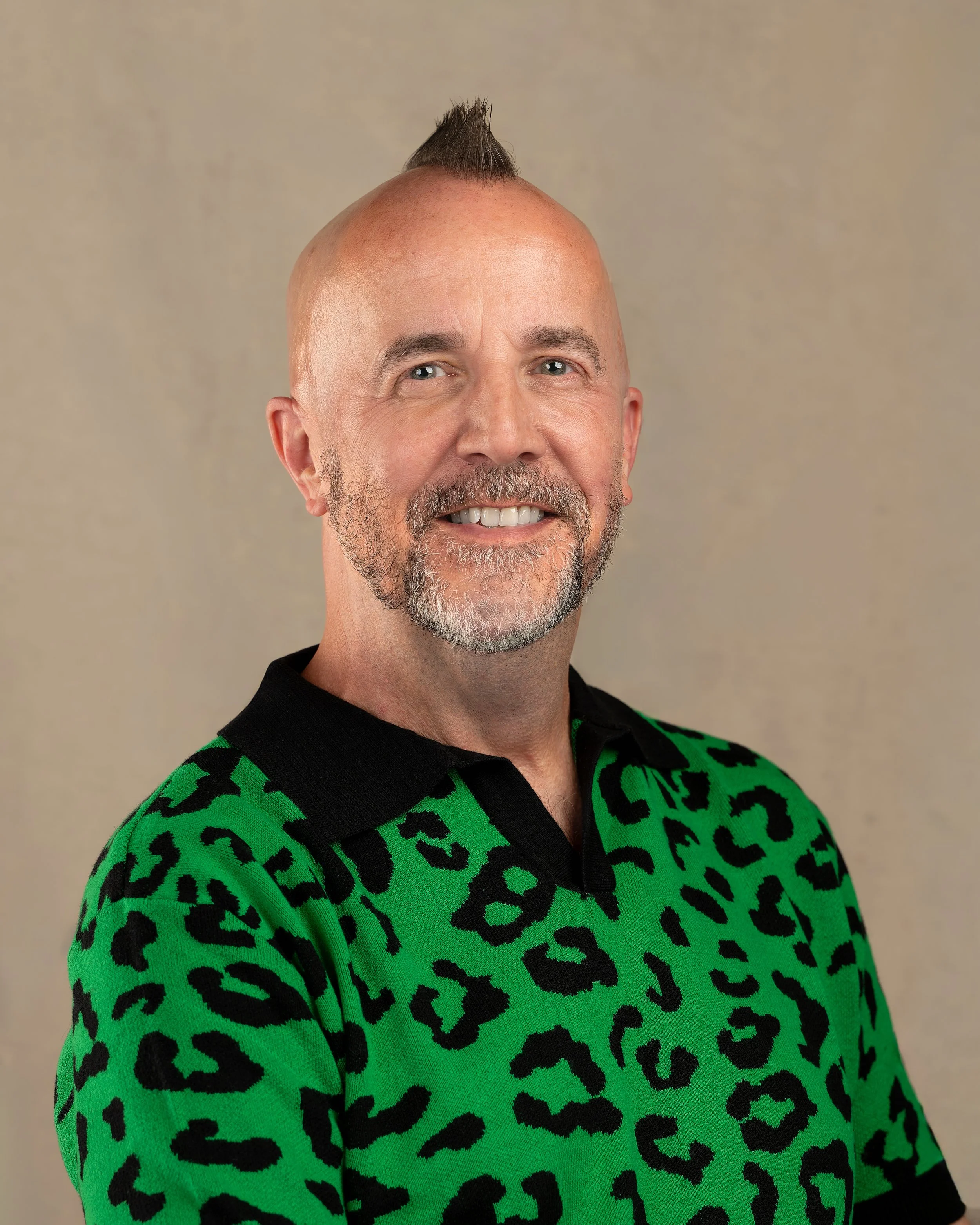 A smiling man with a mohawk hairstyle, beard, and mustache wearing a green sweater with black leopard print and a black collar.