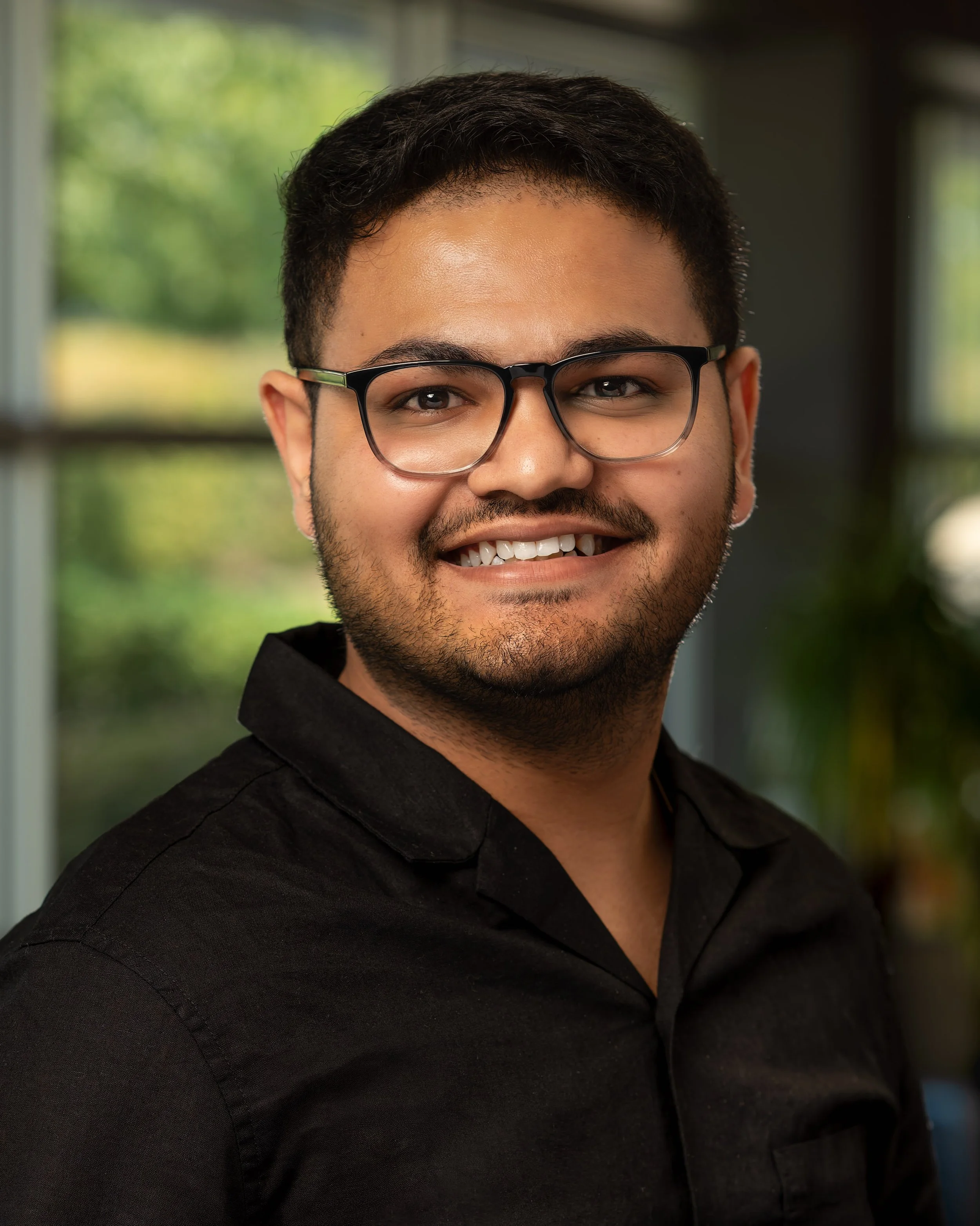 A headshot of young man with glasses smiling in an indoor setting with windows and greenery outside.