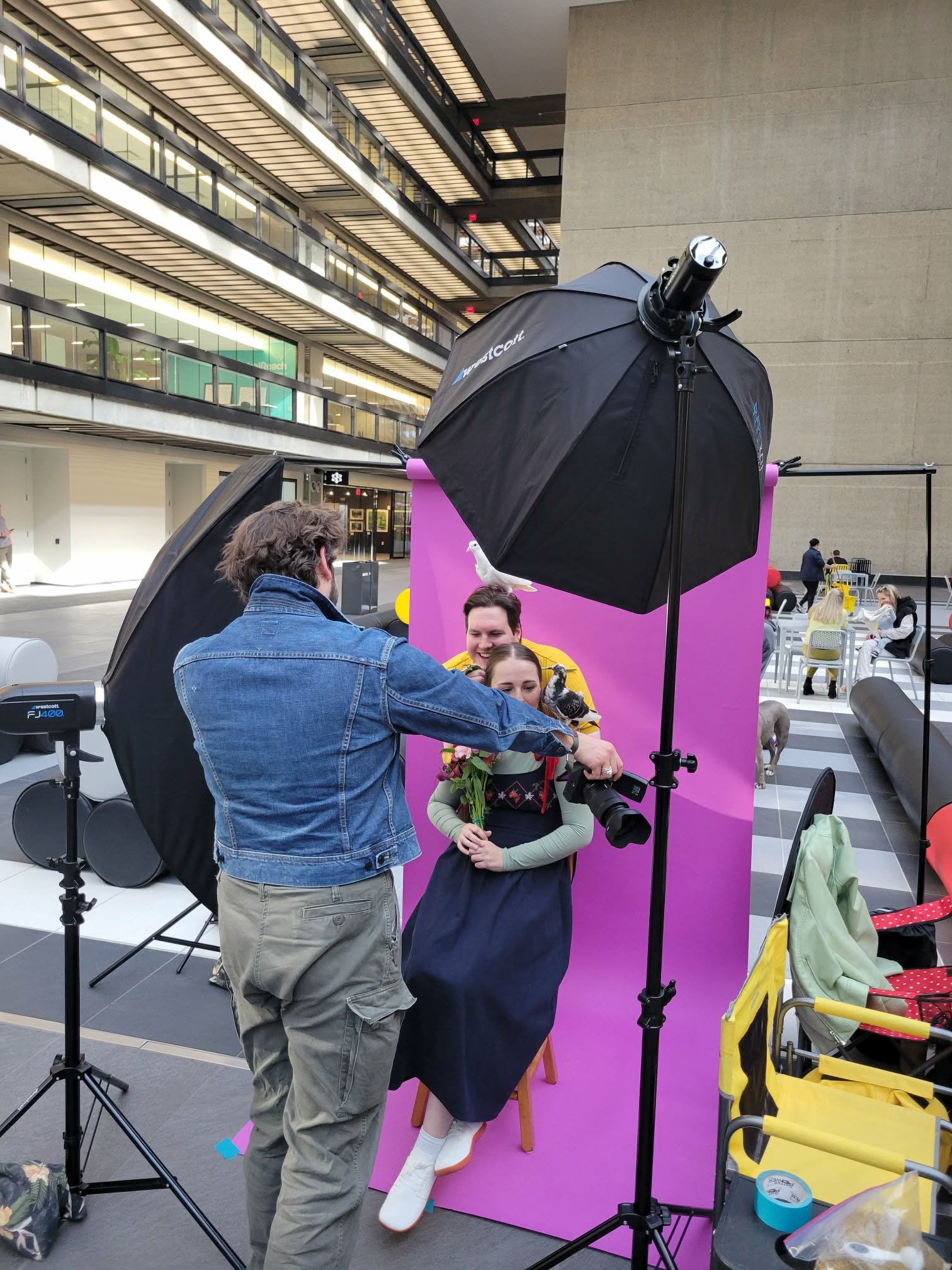 A woman sitting on a stool, holding flowers and a parrot, is being photographed by a man with a camera. The photo is taken in an indoor space with people and seating in the background. Studio lights and umbrellas are set up around the scene.