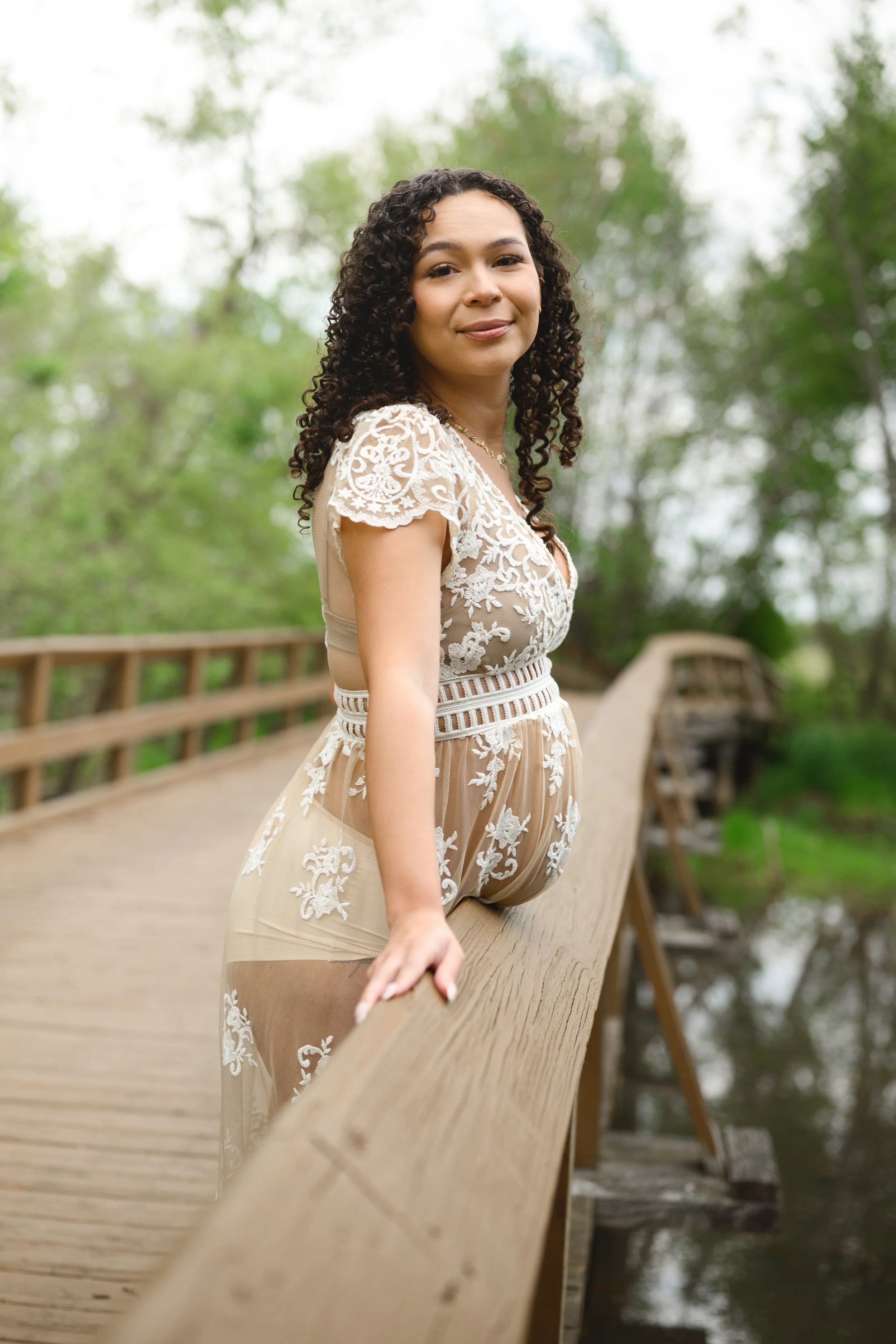 A woman with curly hair wearing a beige embroidered dress standing on a wooden bridge surrounded by trees.