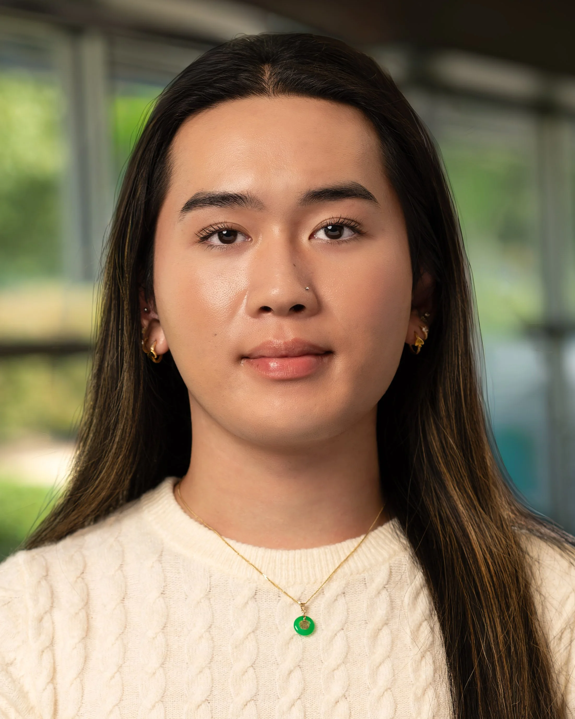 A young woman with long dark hair, wearing a cream-colored sweater and gold jewelry, standing indoors with a blurred window background.