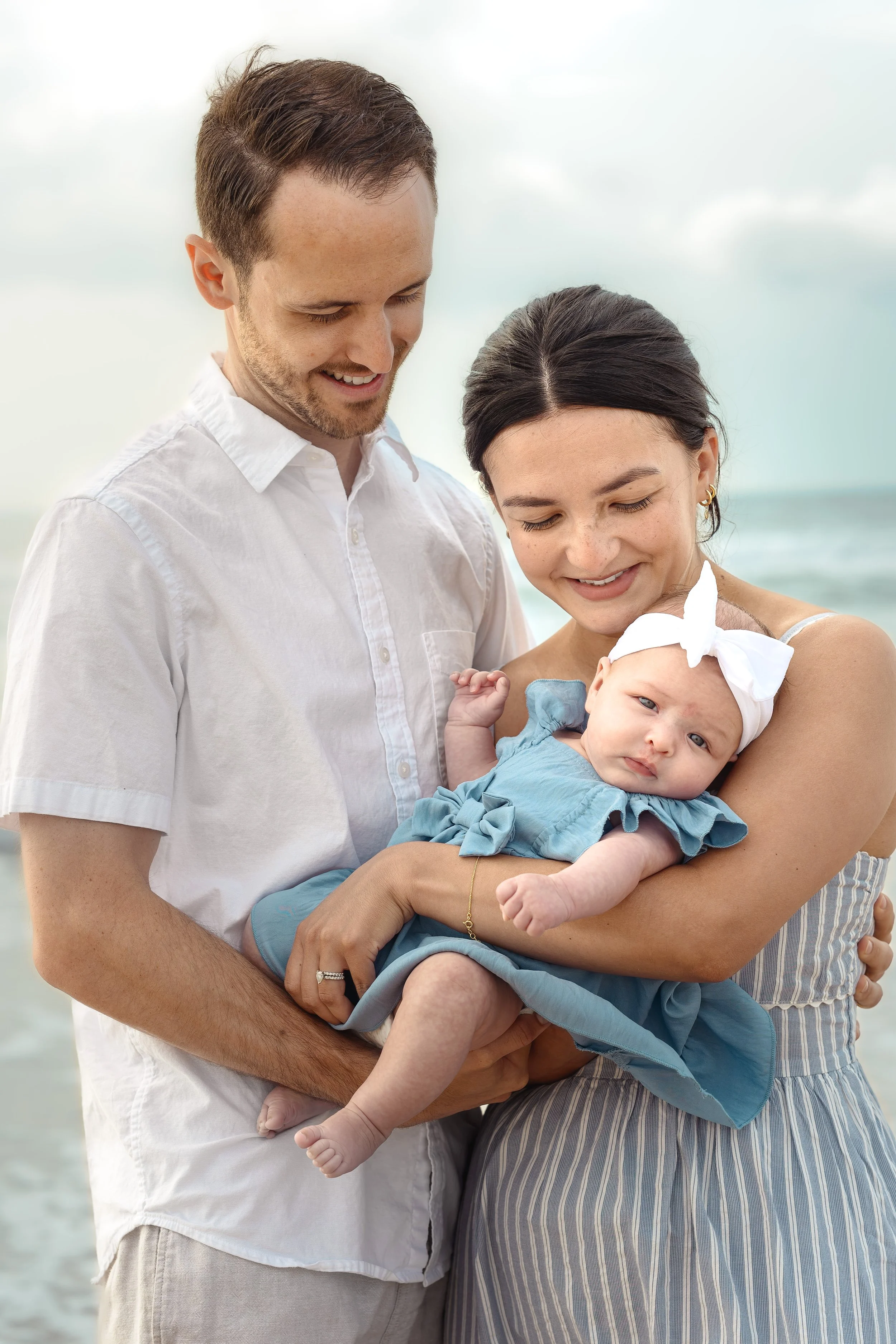 A happy family of three, a man, a woman, and a baby girl, standing on the beach. The man and woman are smiling and looking at their baby, who is dressed in a blue outfit with a white bow on her head.