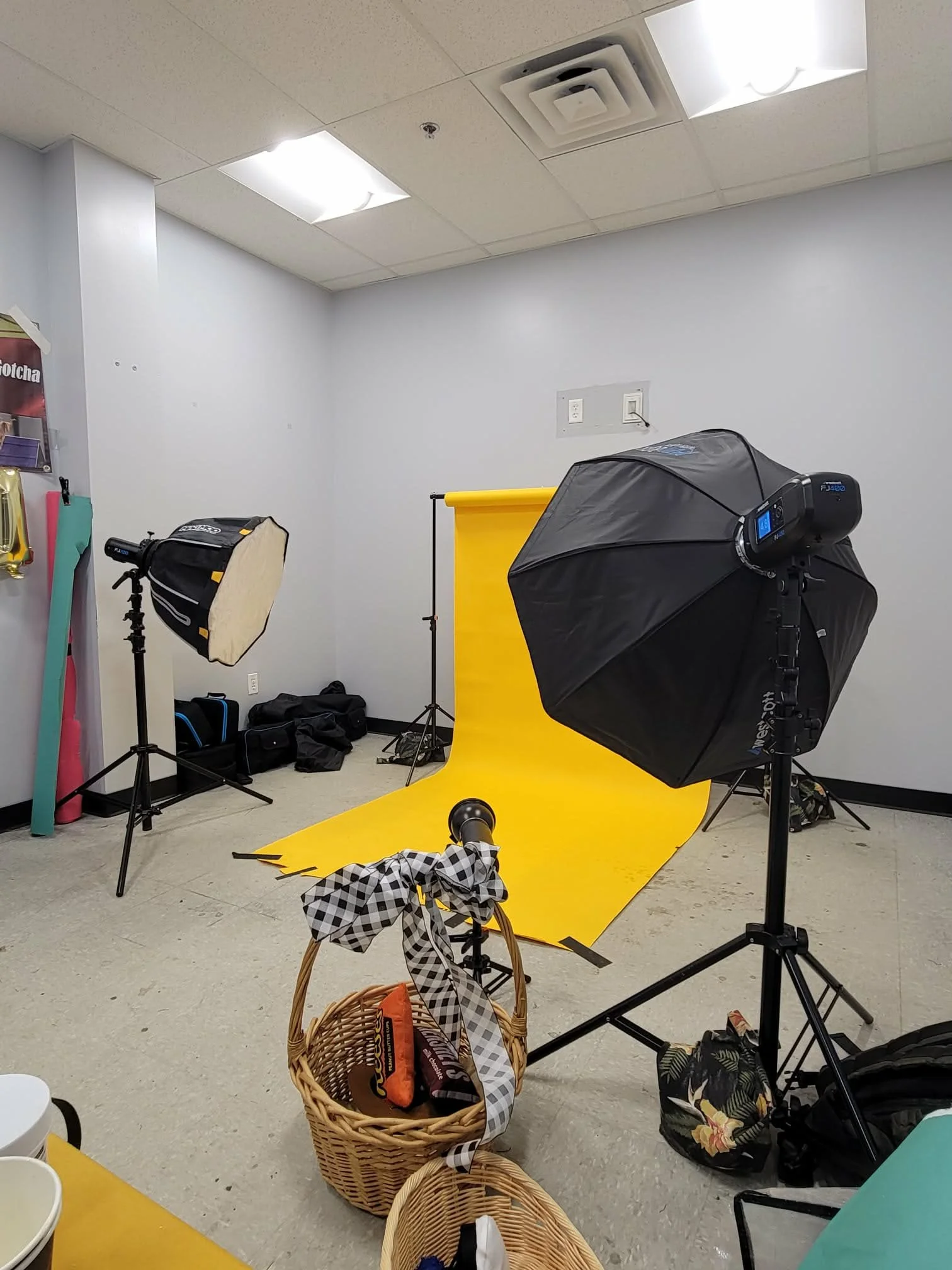Photography studio setup with yellow backdrop, softbox lighting, professional lighting equipment, and photography accessories in a room with plain white walls and ceiling.