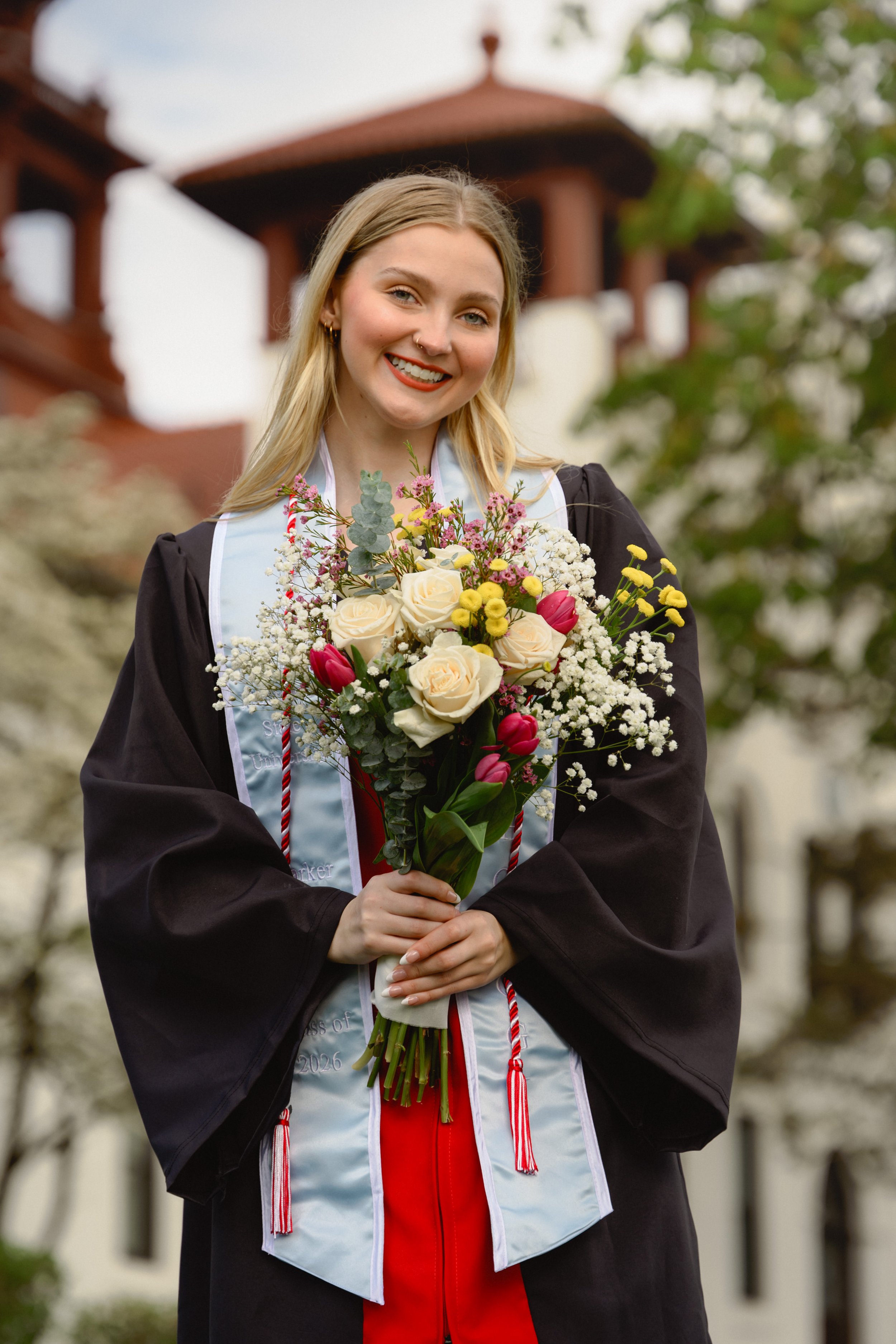A young woman in graduation attire holding a bouquet of flowers outdoors, smiling.