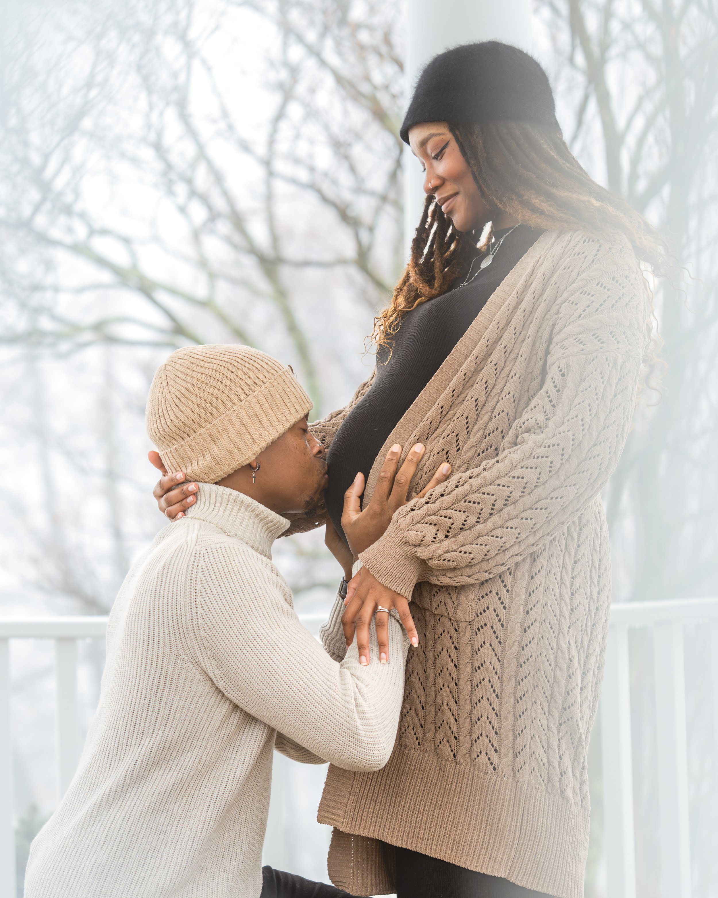 A married pregnant couple outside in the snow during a maternity photoshoot, the husband kneeling and kissing the mother-to-be on the baby bump while she holds him and smiles