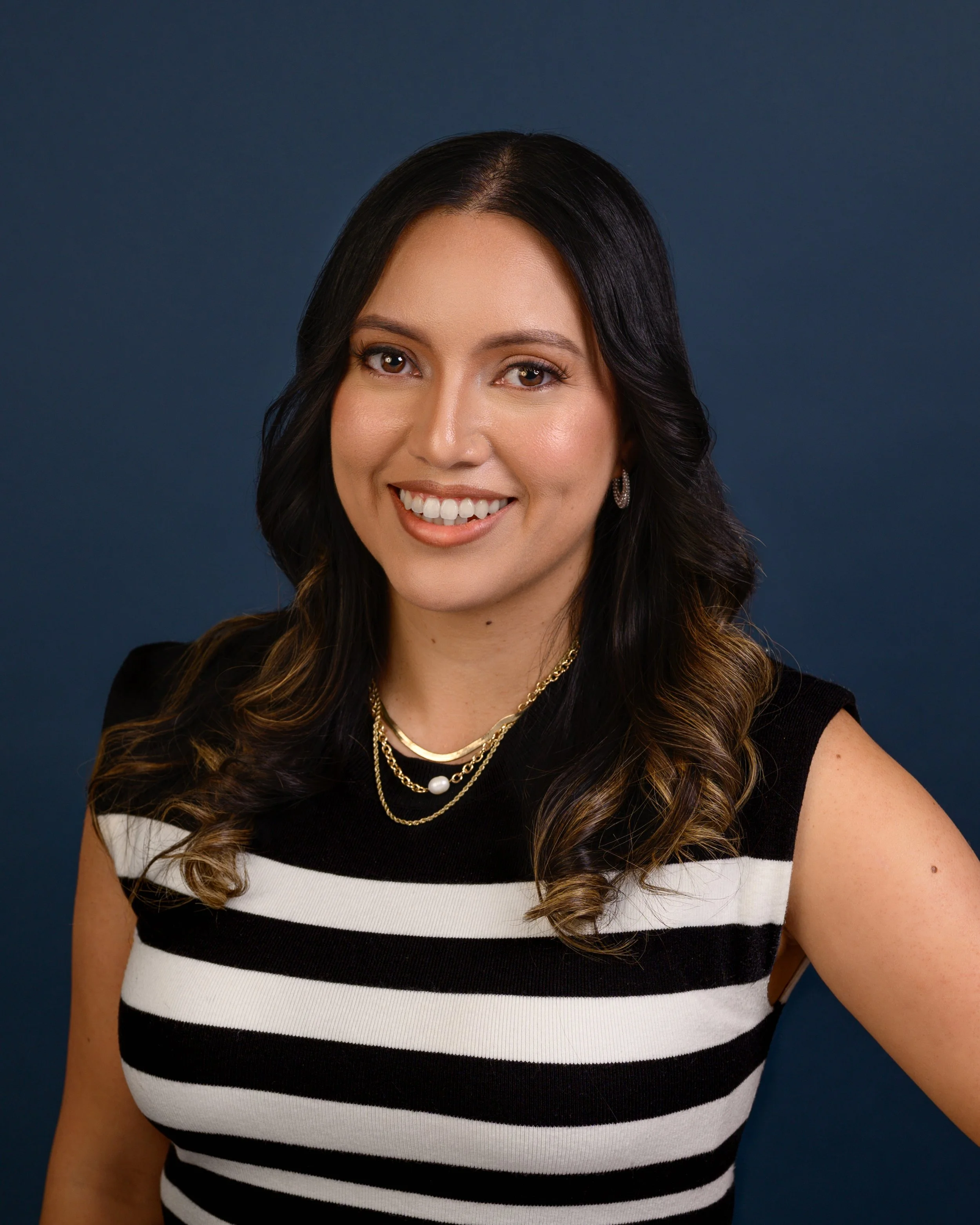 A headshot of a woman with long dark hair and light skin, smiling, wearing a black and white striped sleeveless top and layered gold necklaces, against a plain dark blue background.