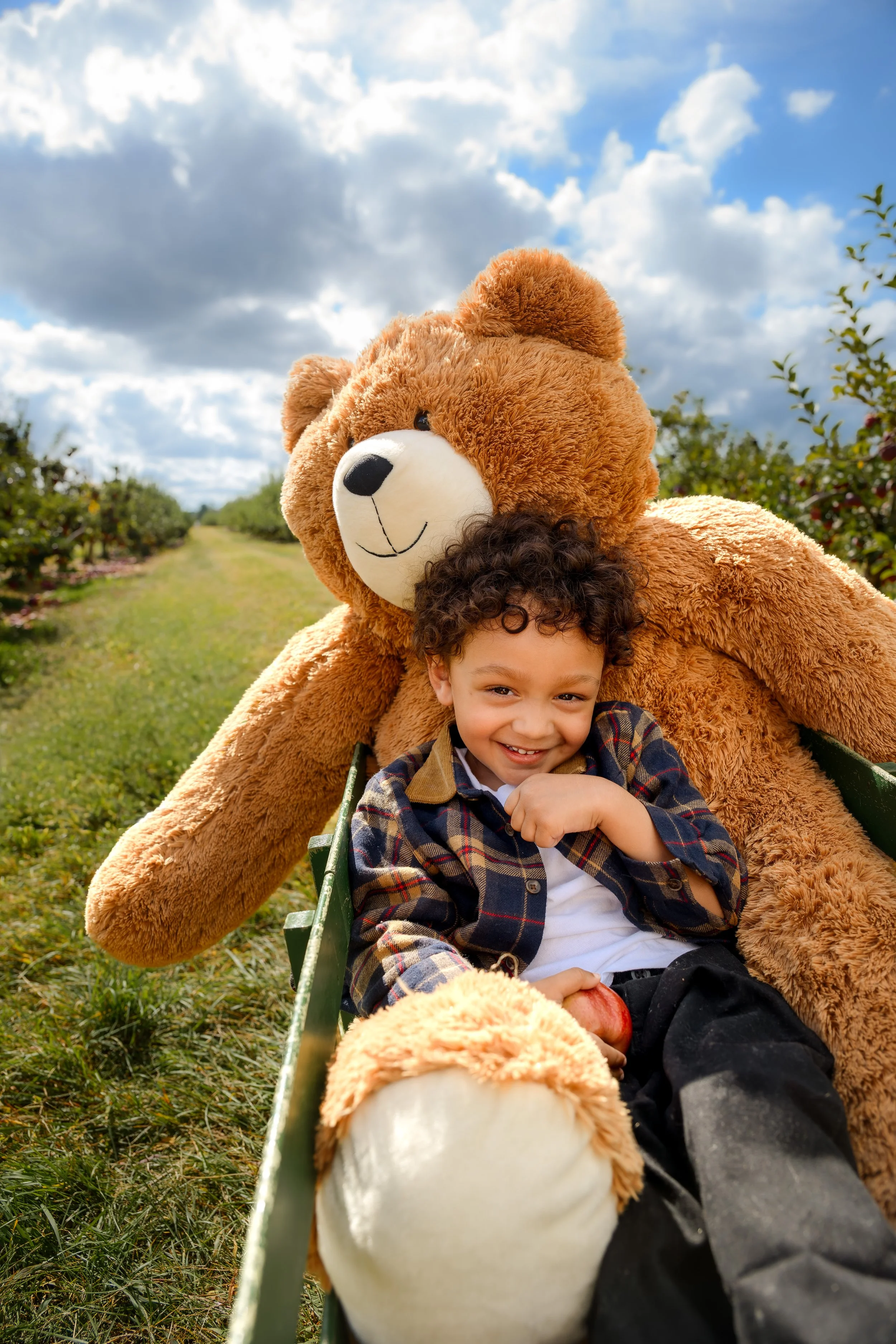 A young boy sitting on a bench with a large teddy bear in an outdoor setting with a cloudy sky.